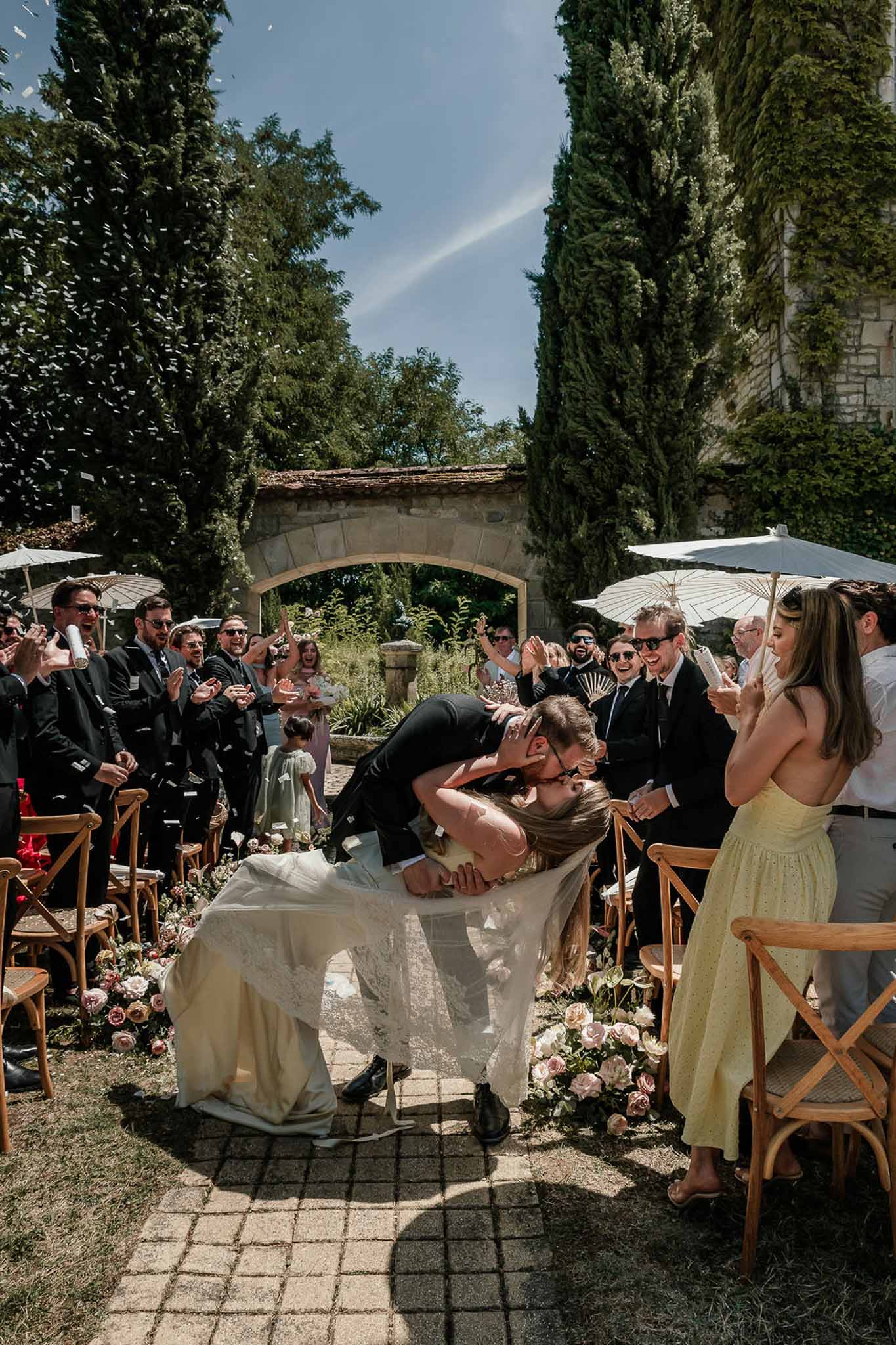 Groom dipping bride for kiss as guests throw confetti in garden courtyard ceremony