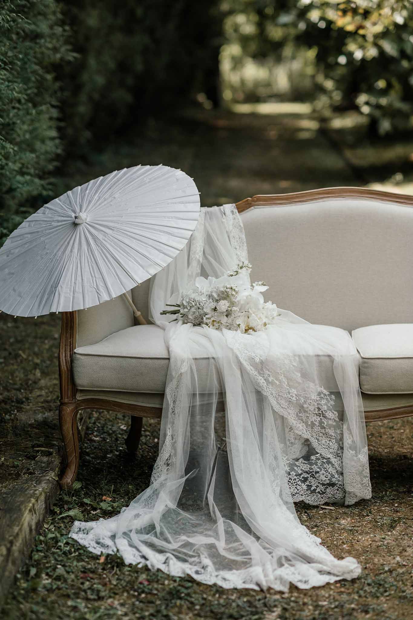 Bridal veil draped over cream Louis XVI settee with white peony bouquet and paper parasol in garden