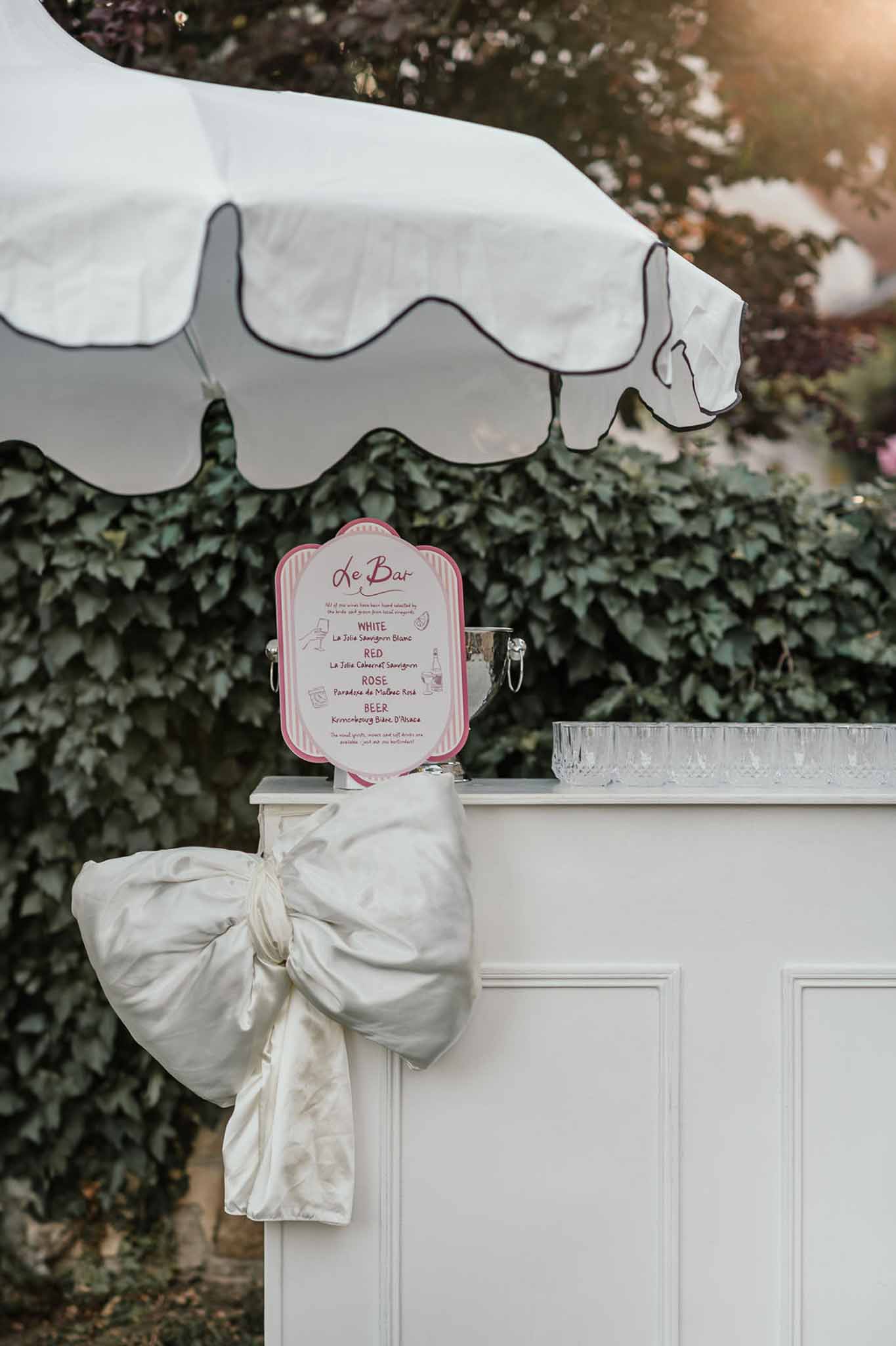 Outdoor cocktail bar with ivory bow, pink and white striped Le Bar menu sign, and crystal glasses