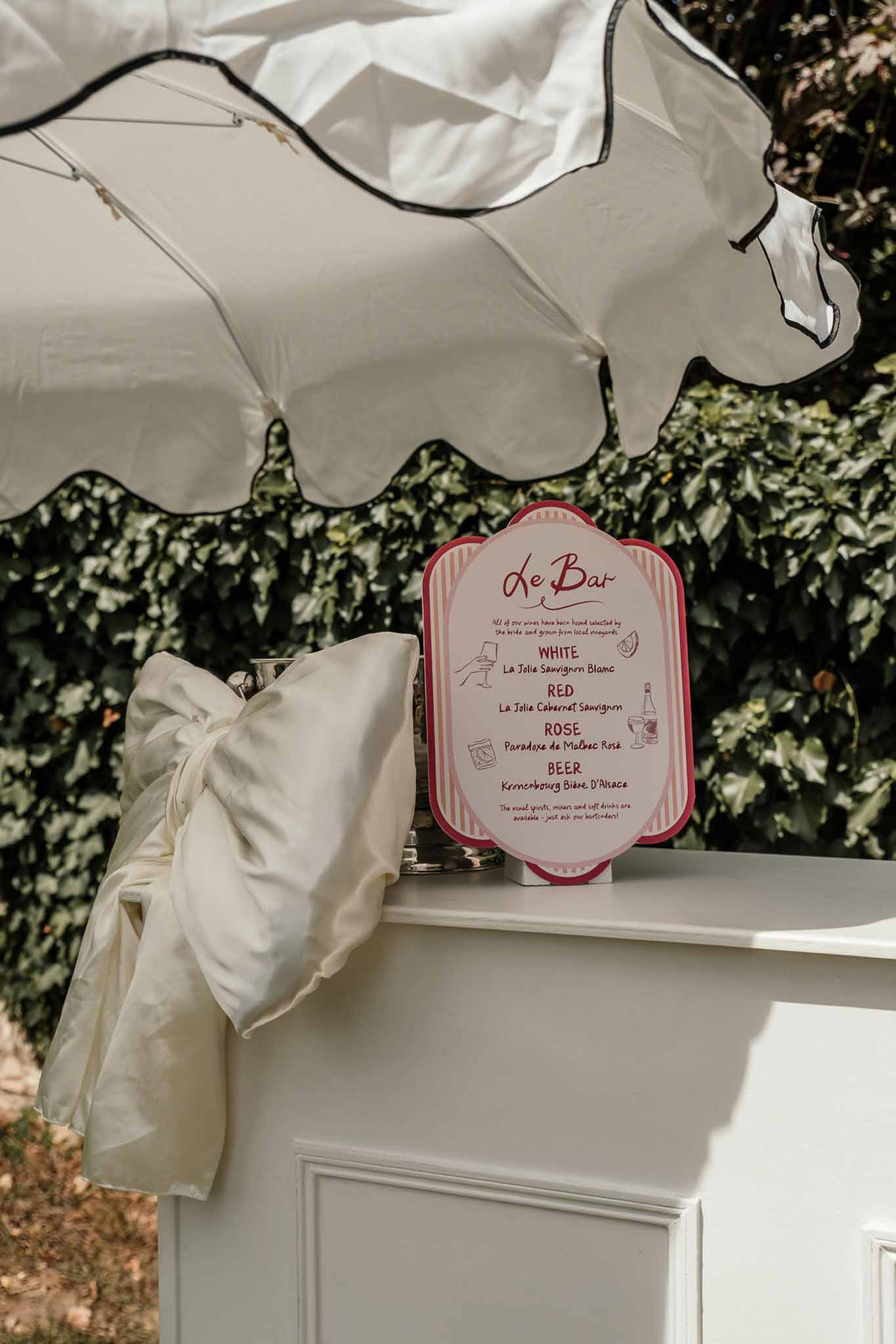 White bar cart with ivory bow and Le Bar menu sign listing wines and beer under scalloped umbrella