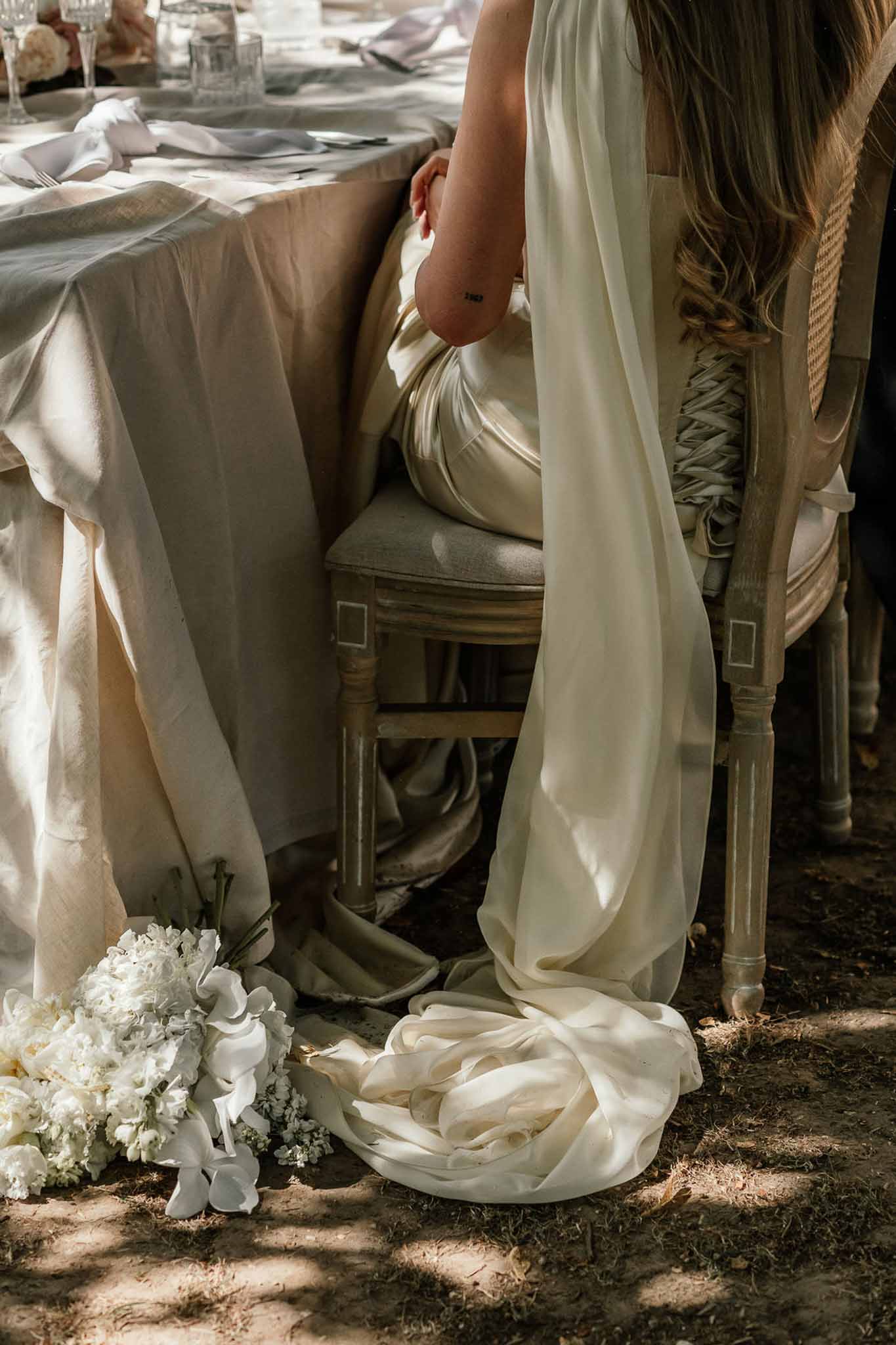 Bride seated at reception table with ivory satin gown pooling on ground beside white sweet pea bouquet