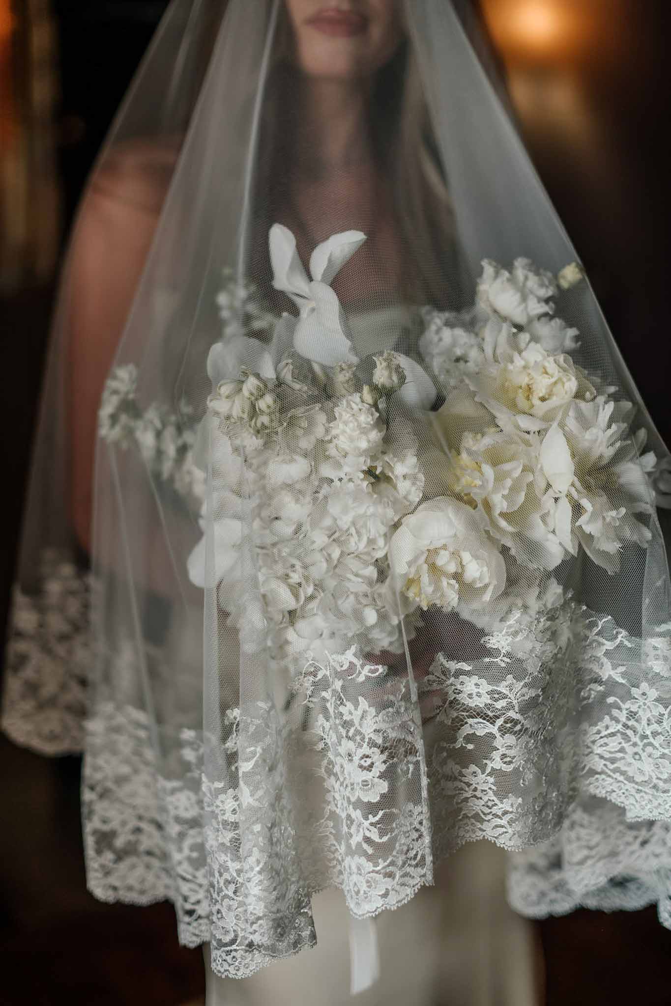 Close-up of bride holding white peony and hydrangea bouquet with floral lace-edged veil draped over flowers