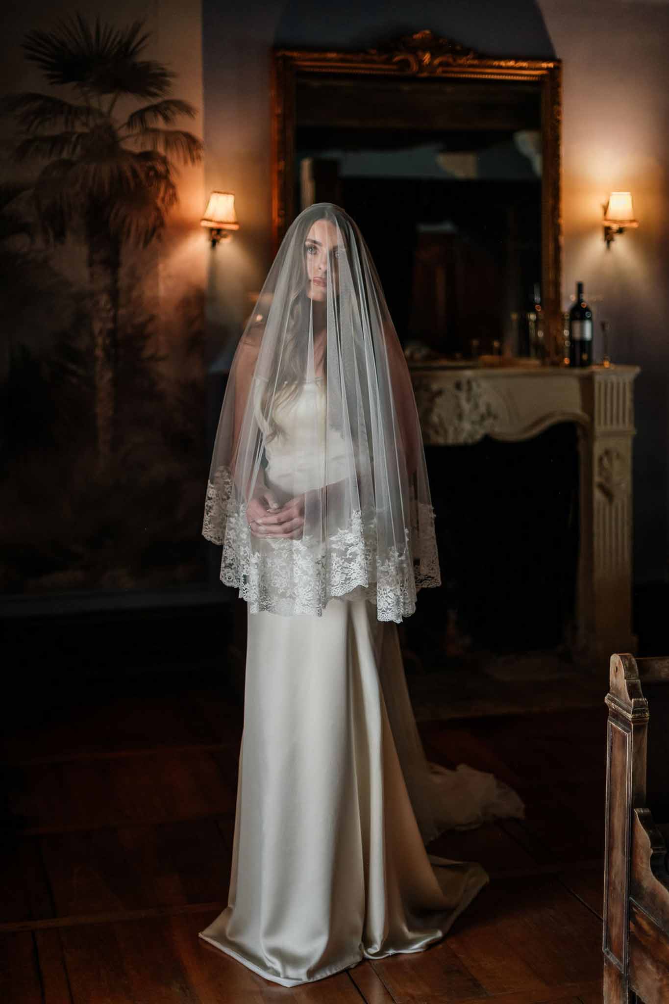 Bride in ivory satin gown with wide lace-bordered veil in dimly lit room with fireplace and wall sconces
