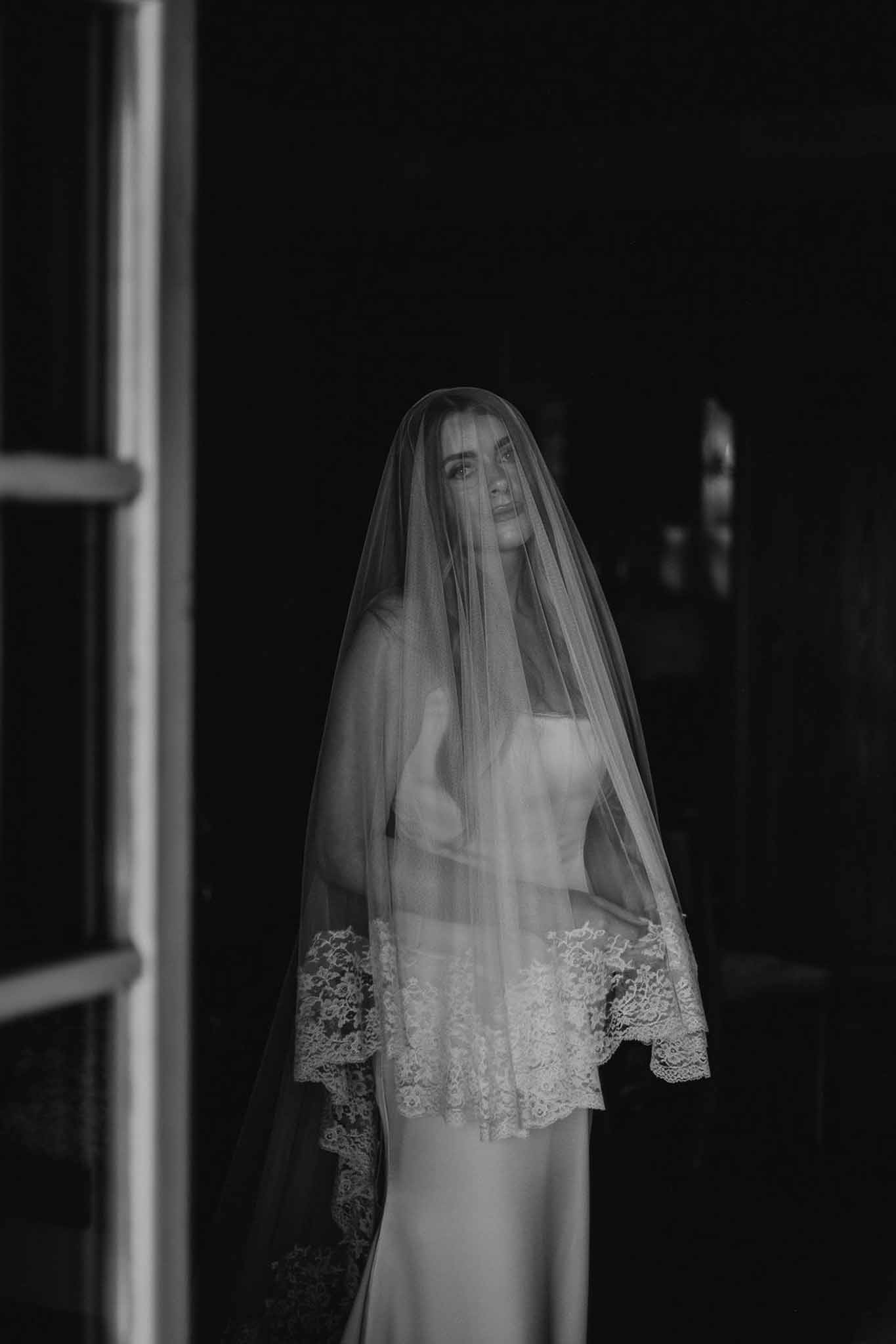 Black and white bride behind floor-length lace-bordered veil gazing upward in dramatic window light