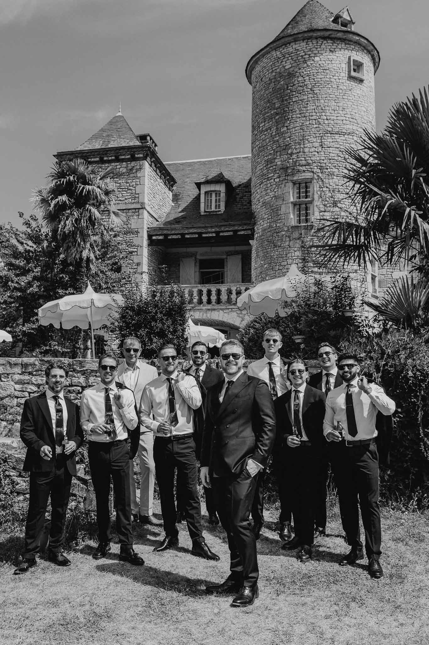 Black and white group portrait of groom and groomsmen with drinks and cigars outside chateau with round tower