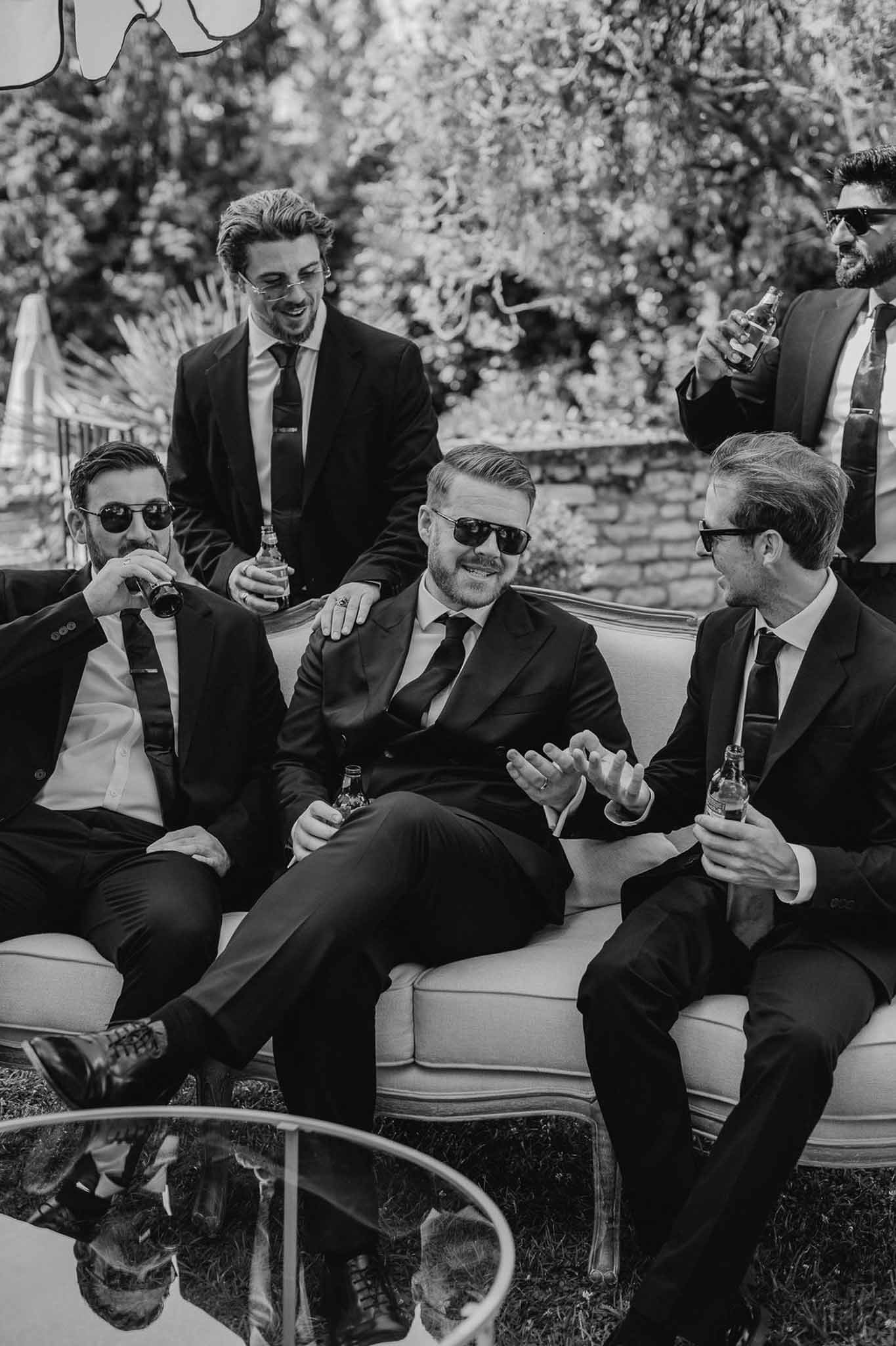 Black and white five groomsmen in dark suits with beers on vintage sofa during outdoor cocktail hour