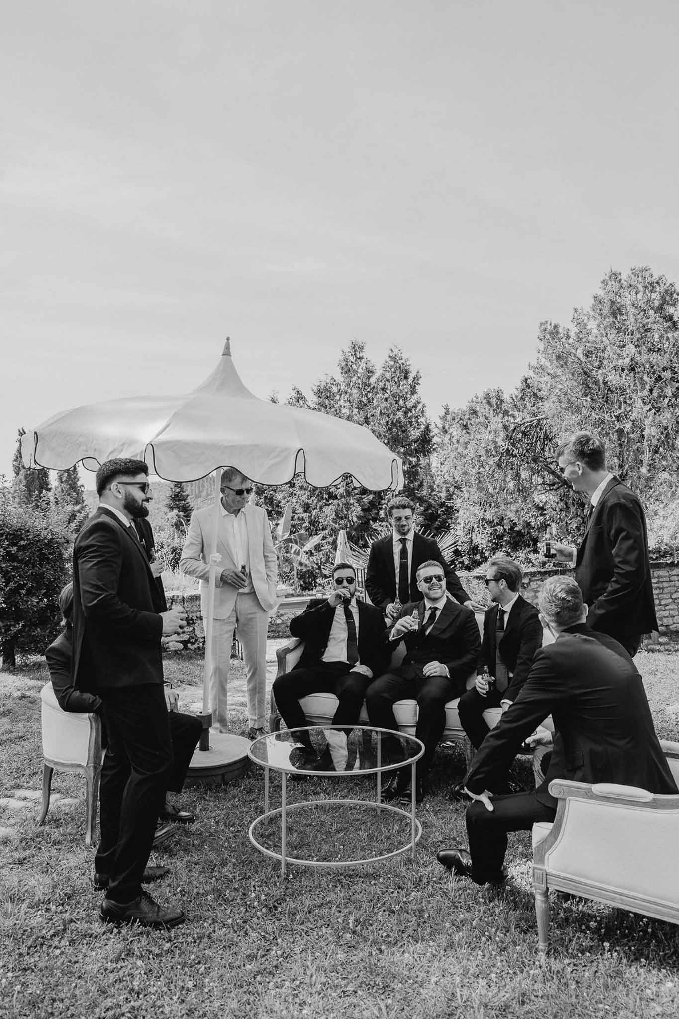 Group of groomsmen in dark suits gathered around lounge furniture under parasol during outdoor cocktail hour