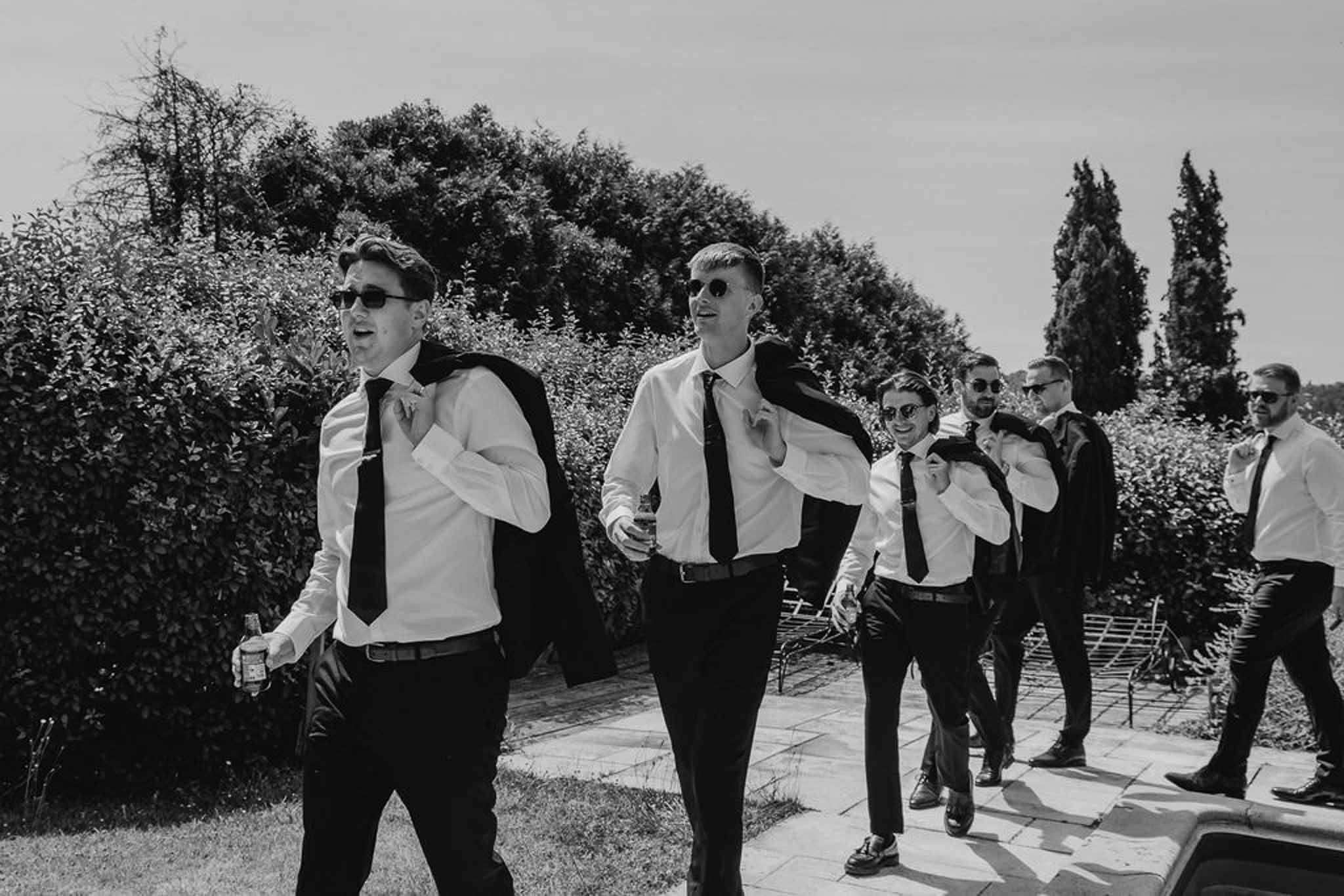 Six groomsmen walking along garden path carrying jackets over shoulders in black and white