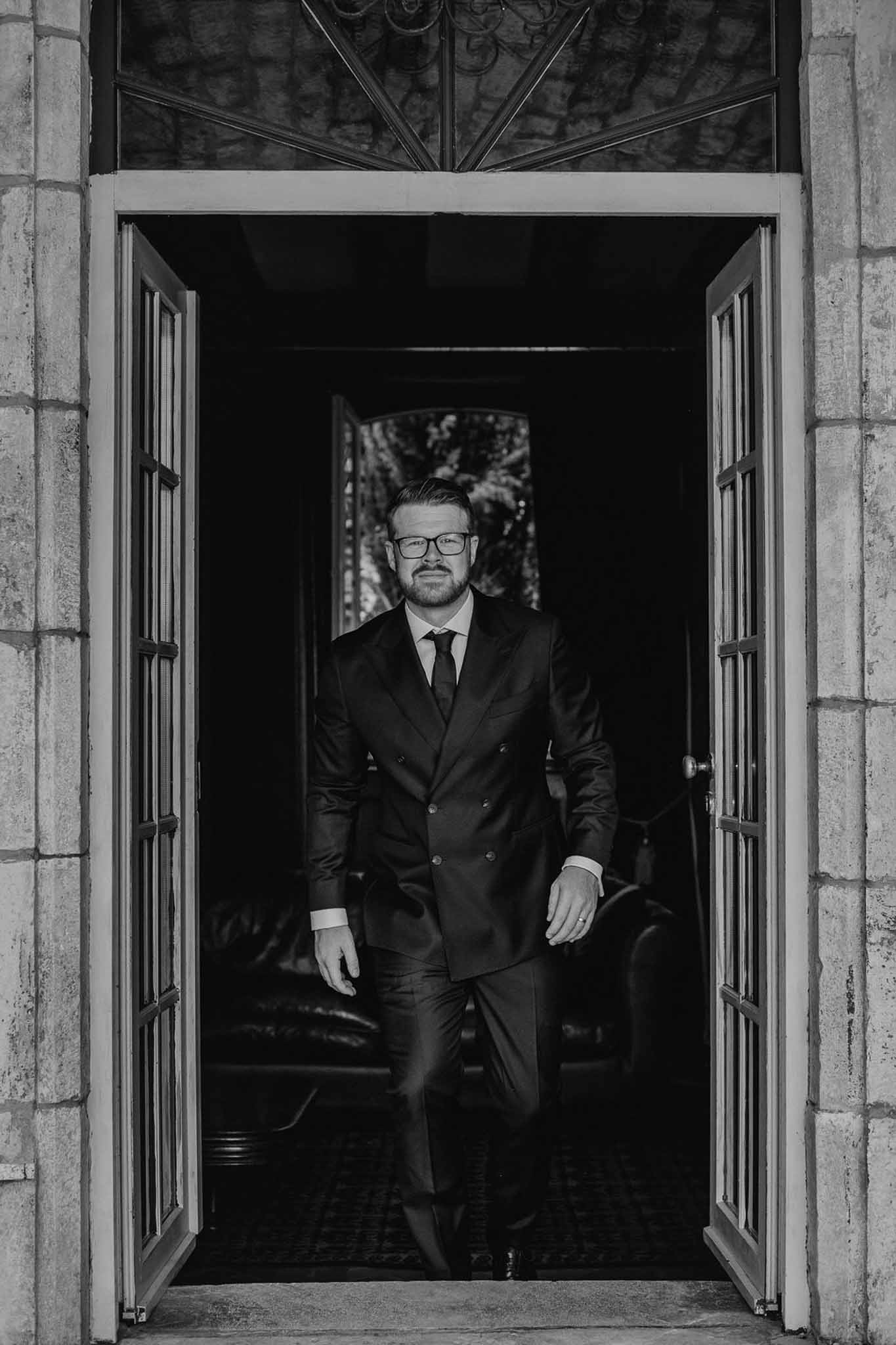 Bearded groom in double-breasted suit walks through stone doorway with wrought-iron fanlight in B&W