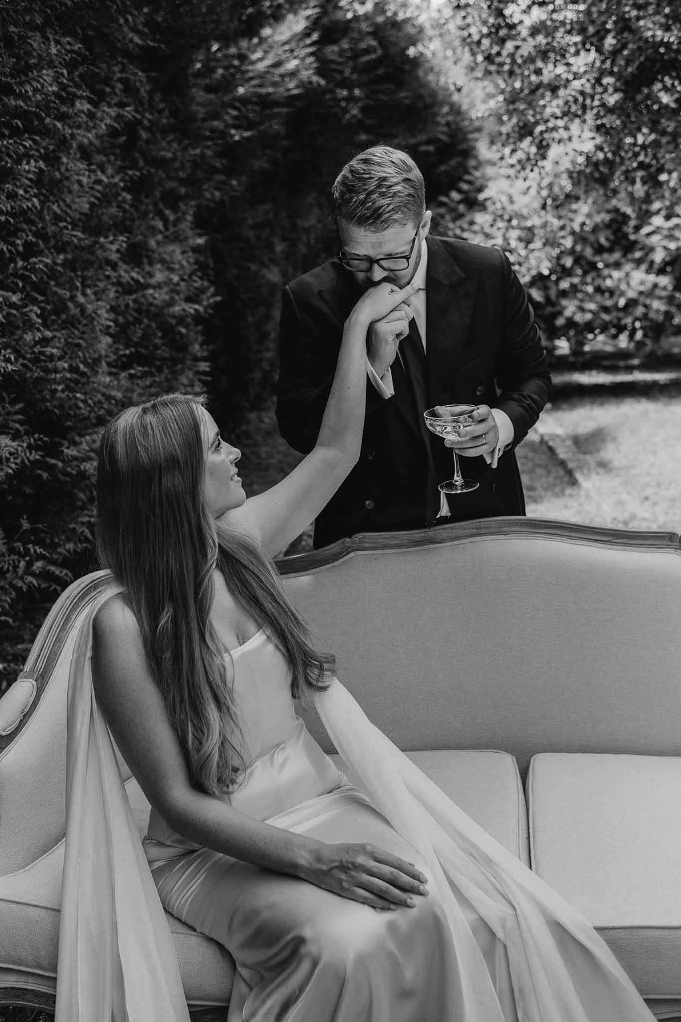 Black and white portrait of groom kissing bride's hand while she reclines on vintage outdoor sofa