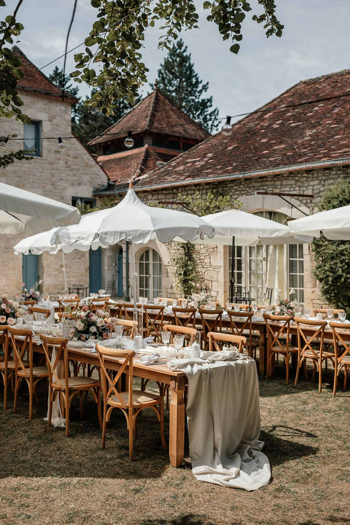 Farm tables with sage runners and blush rose centerpieces under white scalloped umbrellas at stone courtyard venue