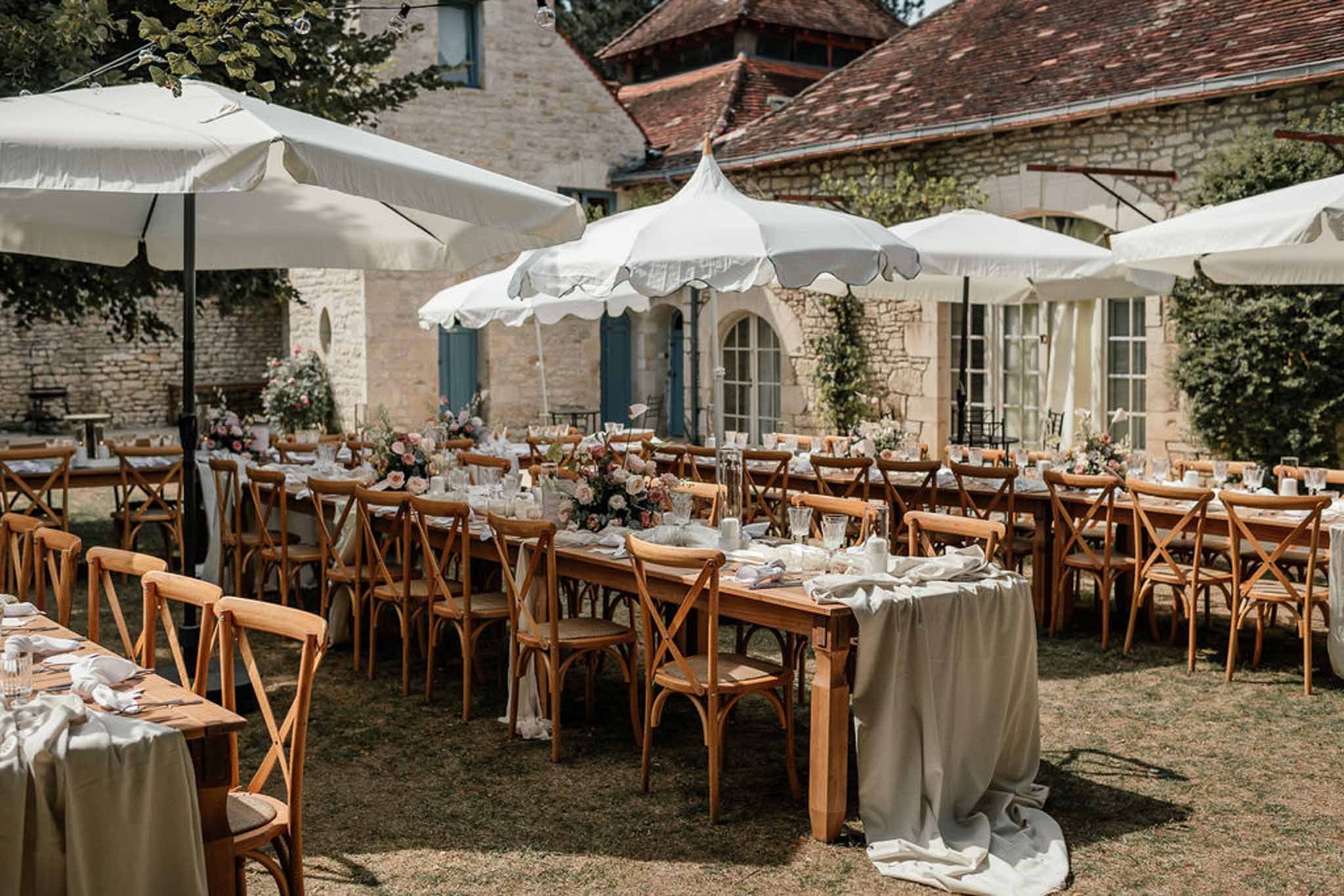 Farm tables with sage linen and blush rose runners under white scalloped umbrellas in stone courtyard