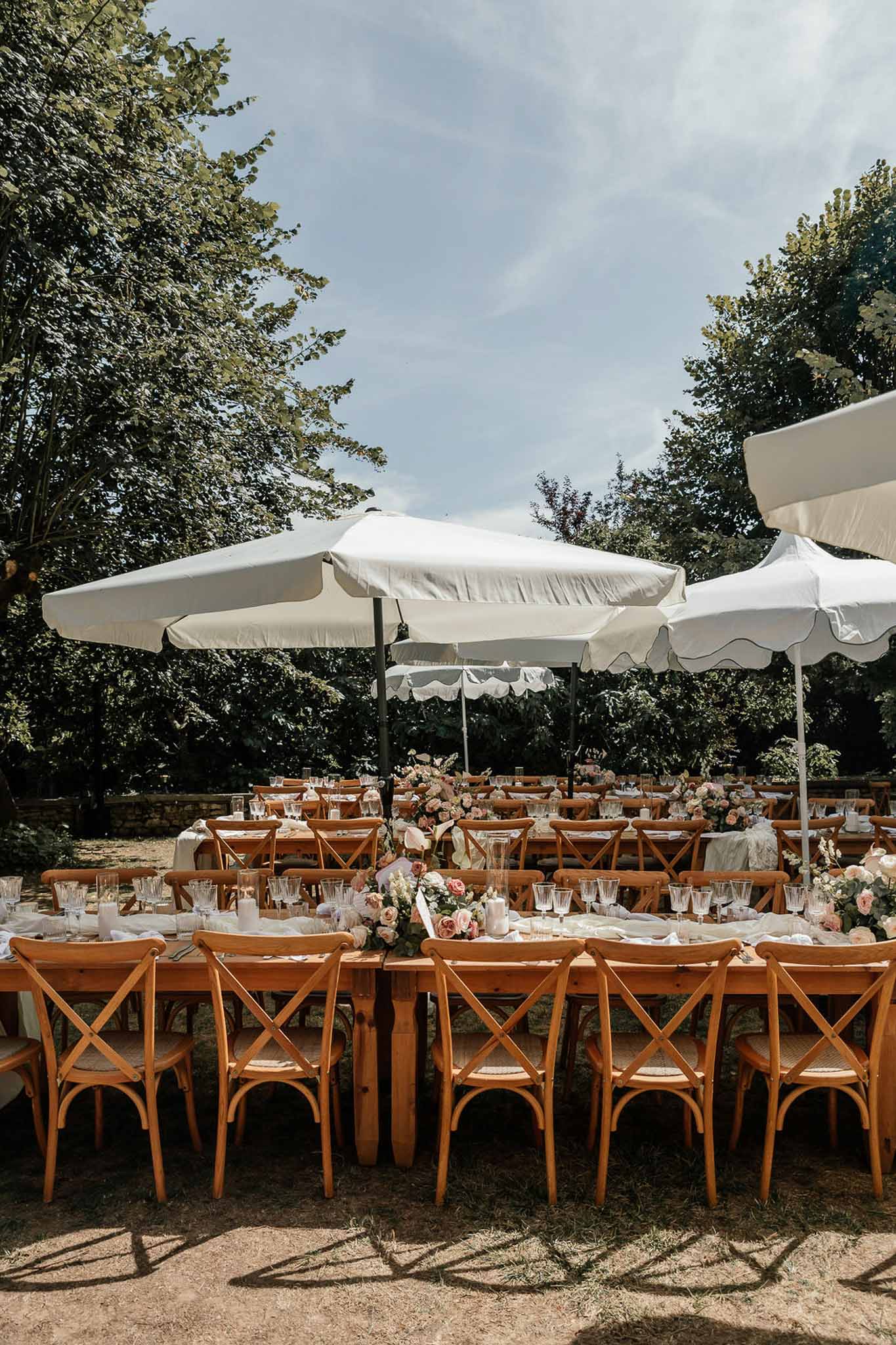 Farm tables with cross-back chairs, blush rose centerpieces, and white scalloped market umbrellas before guests arrive