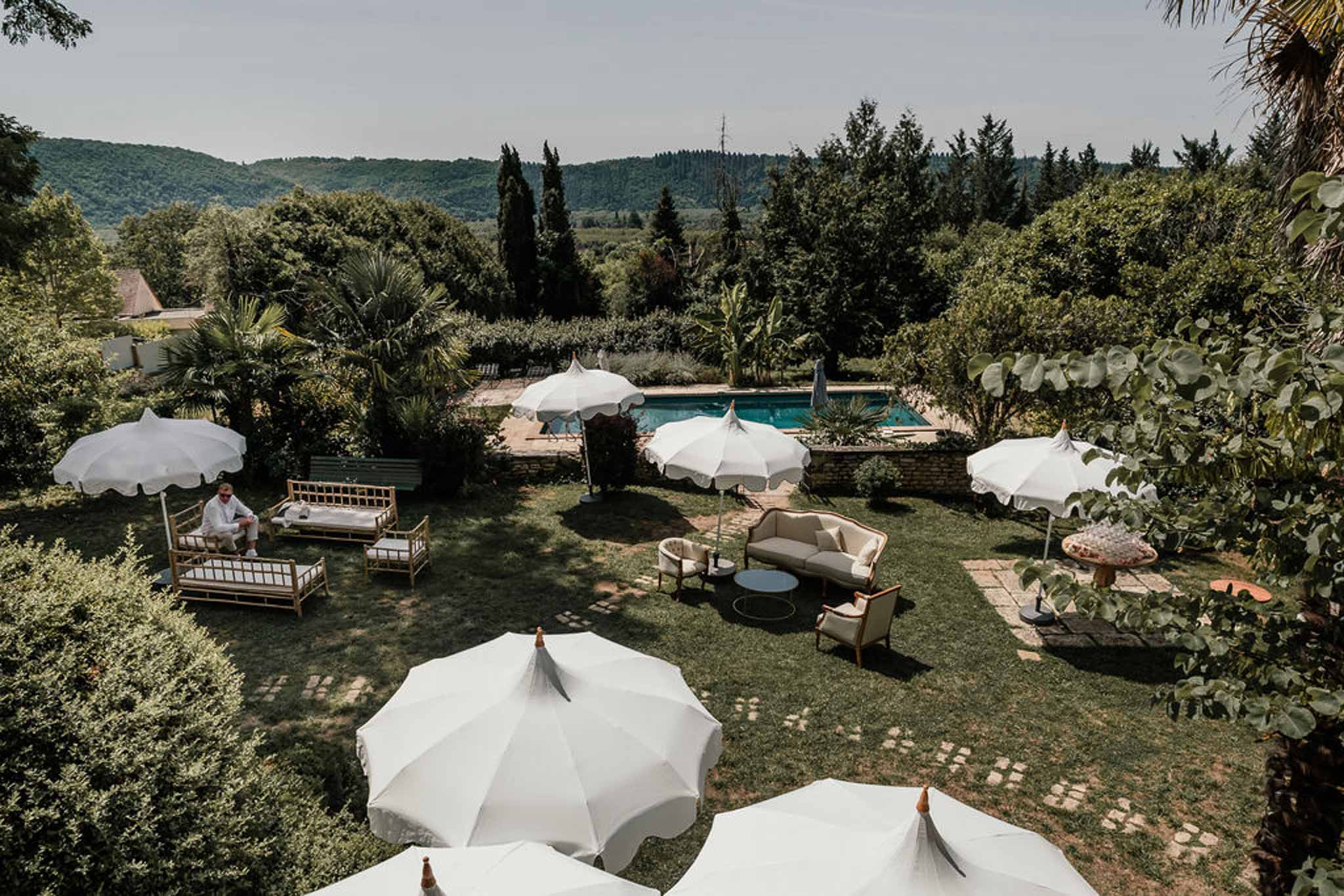 Garden lounge area with bamboo daybeds, white parasols, and cream sofas beside pool and wooded hills