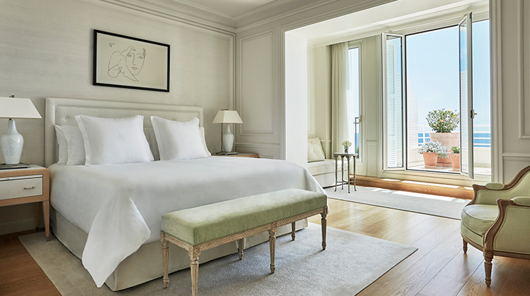 Hotel bedroom suite with king bed, sage green bench, armchair, and French doors opening to sea view terrace