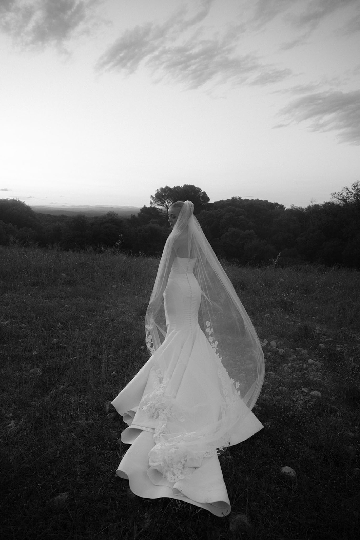 Black and white rear view of bride in mermaid gown and lace-trimmed cathedral veil standing in open field at dusk