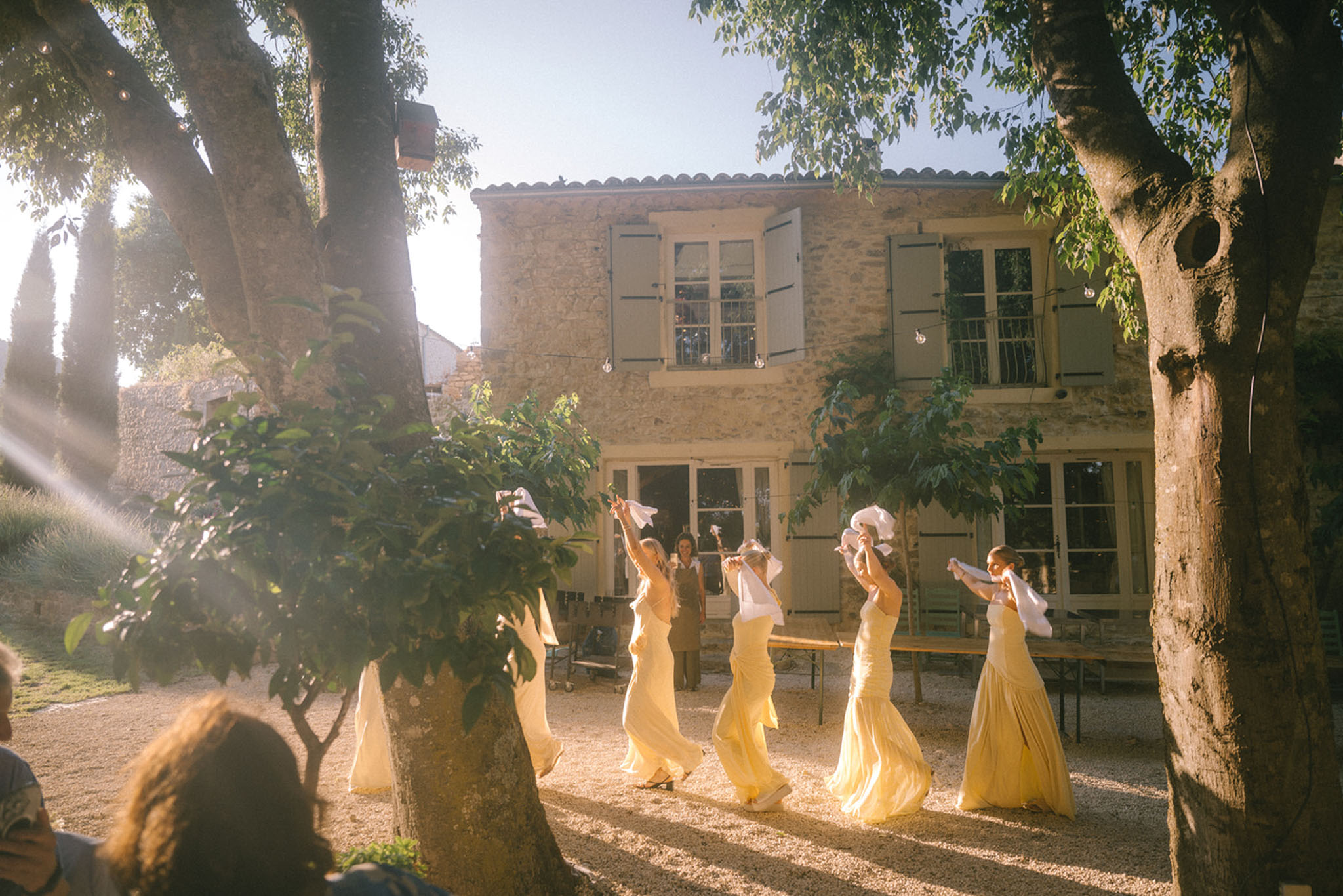 Four bridesmaids in pale yellow chiffon gowns waving white cloths across gravel courtyard at golden hour