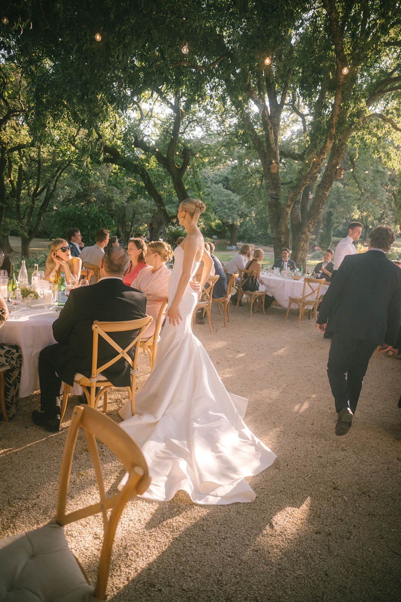 Bride greeting guests at long tables under tree canopy with festoon lights and dappled sunlight