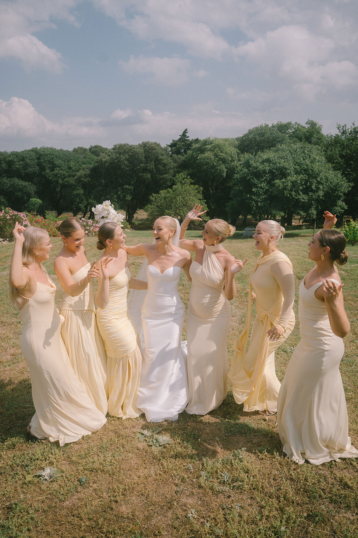 Bride laughing with six bridesmaids in mismatched butter yellow floor-length gowns on sunny garden lawn