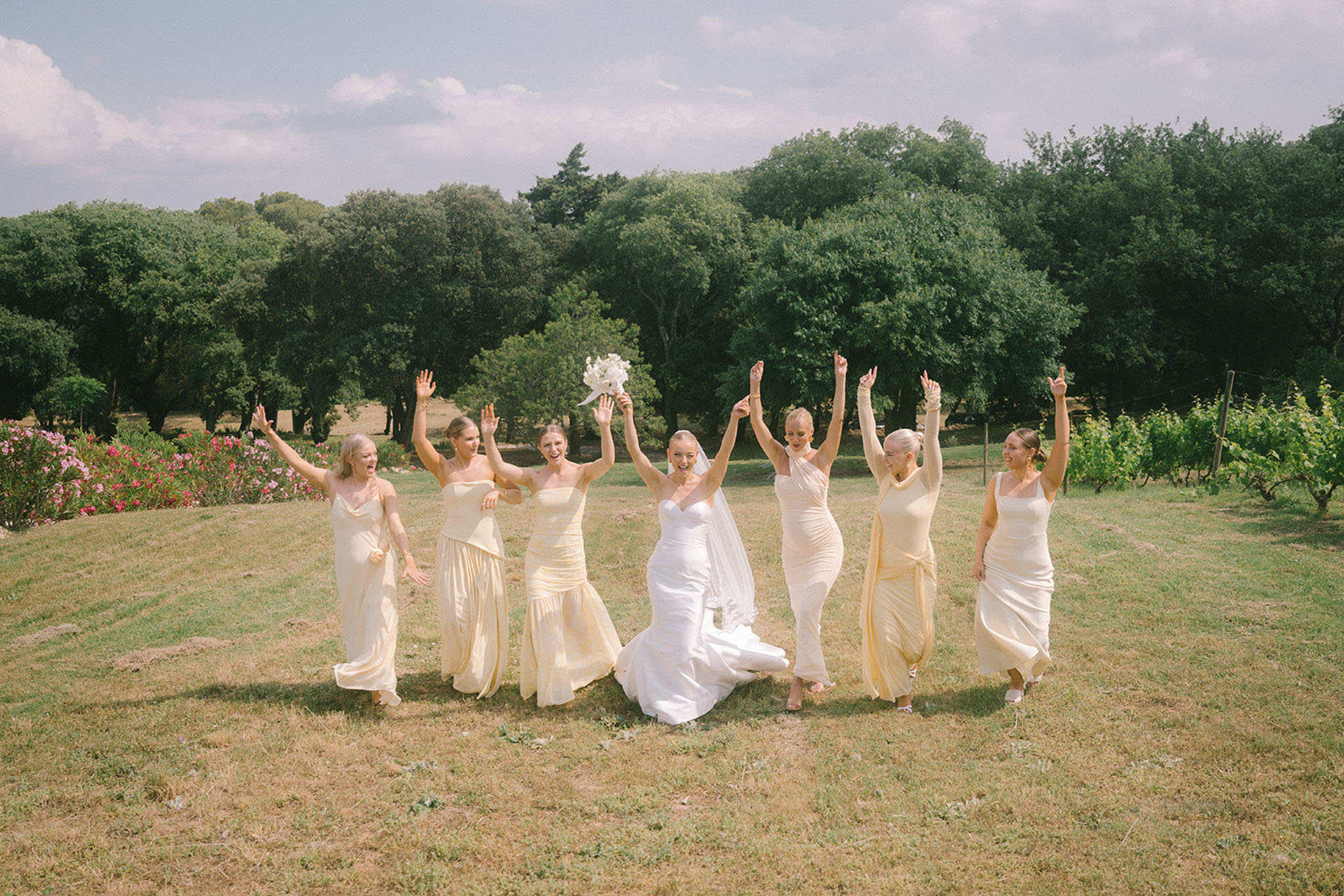 Bride in white strapless gown with six bridesmaids in yellow satin dresses jumping on vineyard estate grounds