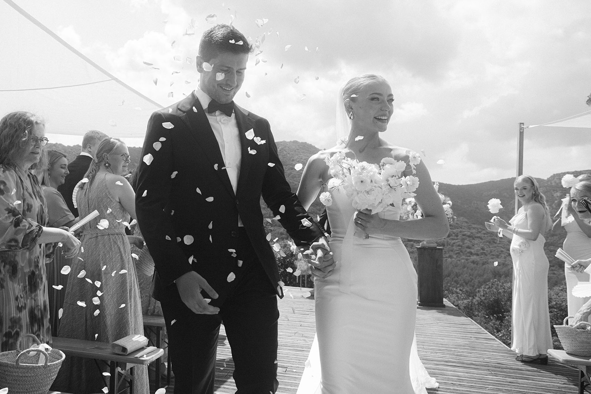 Bride and groom recessional with rose petals tossed on wooden deck with hillside backdrop in B&W
