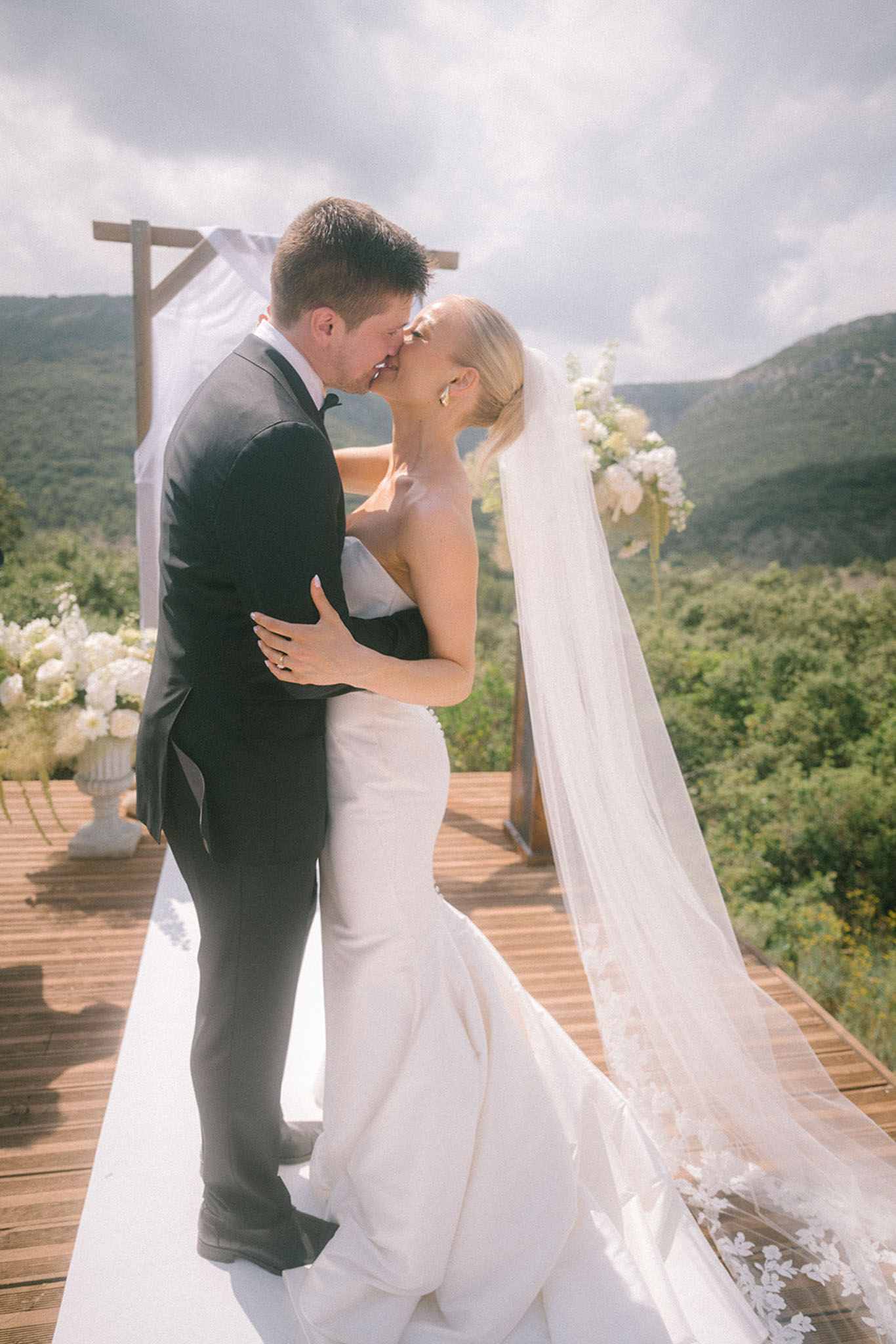 Bride and groom share first kiss under wooden ceremony arch draped in white fabric with mountain valley panorama behind