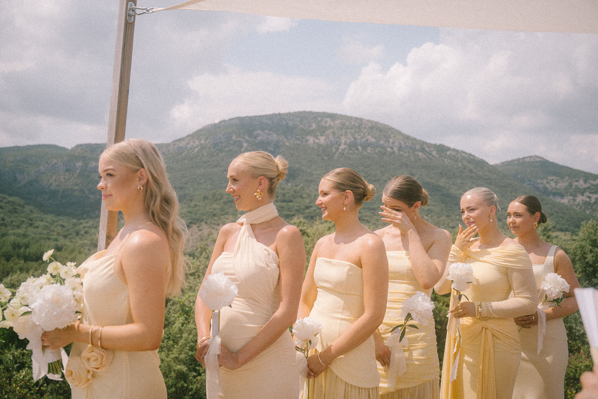 Six emotional bridesmaids in butter yellow and cream dresses holding white peony bouquets at outdoor ceremony