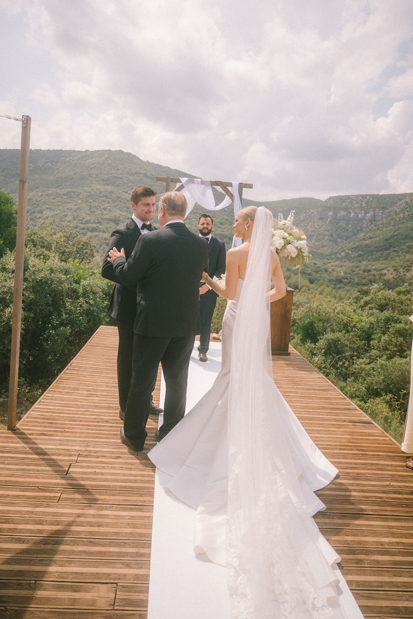 Bride with cathedral veil walked down aisle on mountain-view wooden deck toward emotional groom at arch