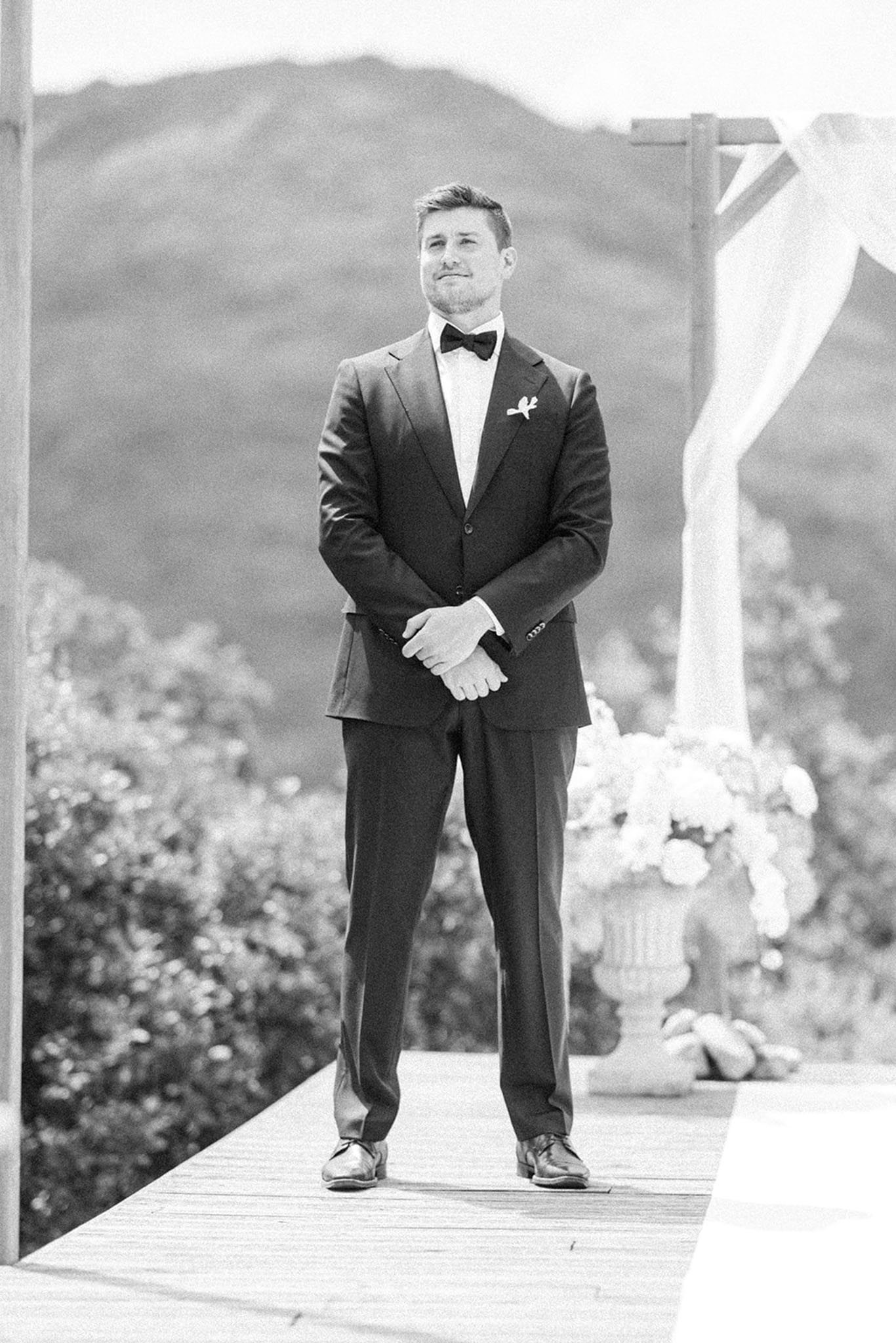 Black and white portrait of groom waiting at outdoor altar with draped arch and urn florals