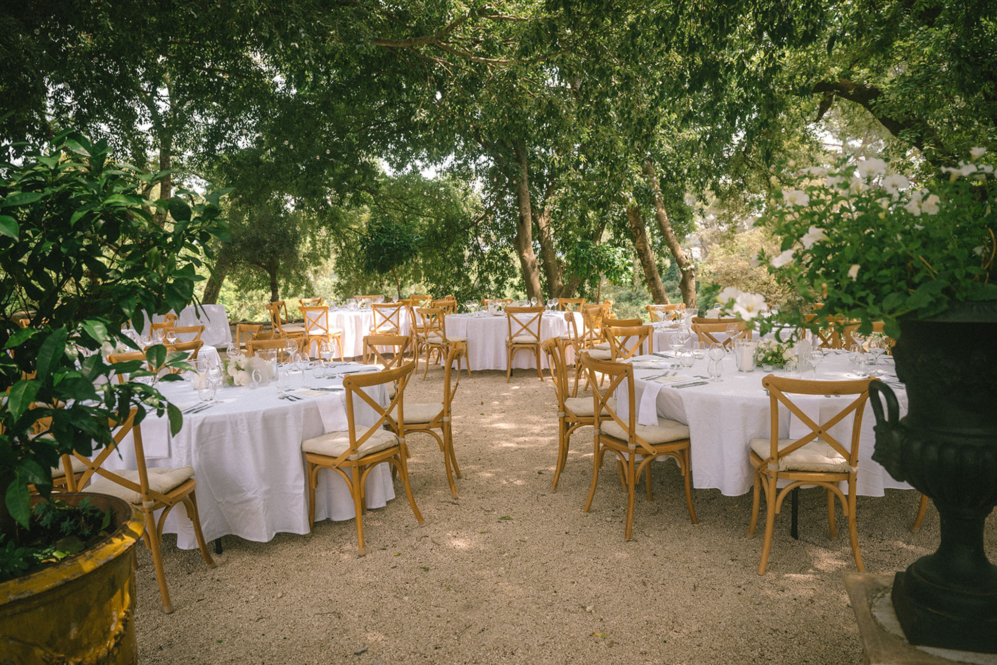 Round reception tables with white linens and cross-back chairs under mature trees in shaded garden