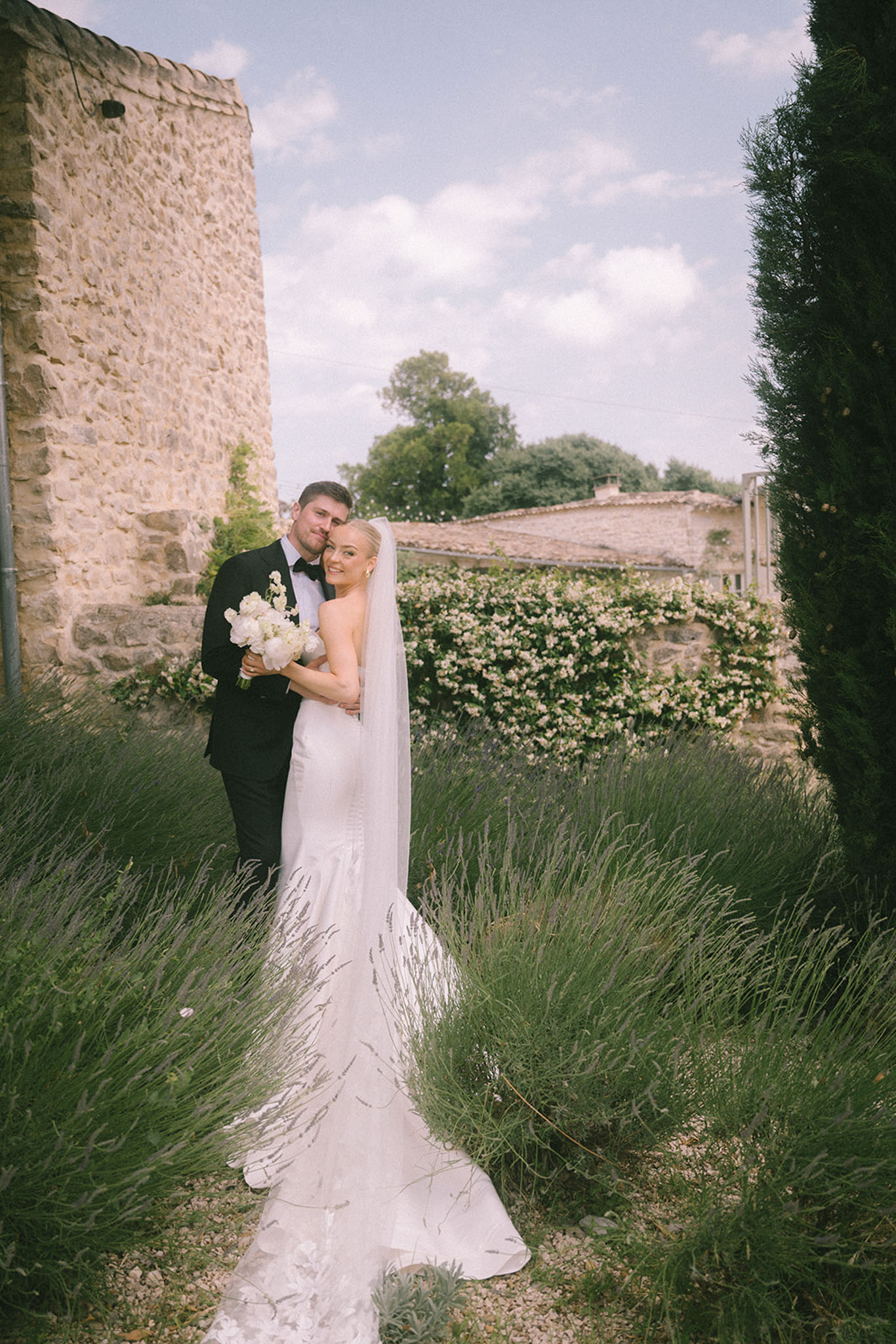Bride in ivory mermaid gown with cathedral veil and groom in black tuxedo among lavender rows at Provencal property