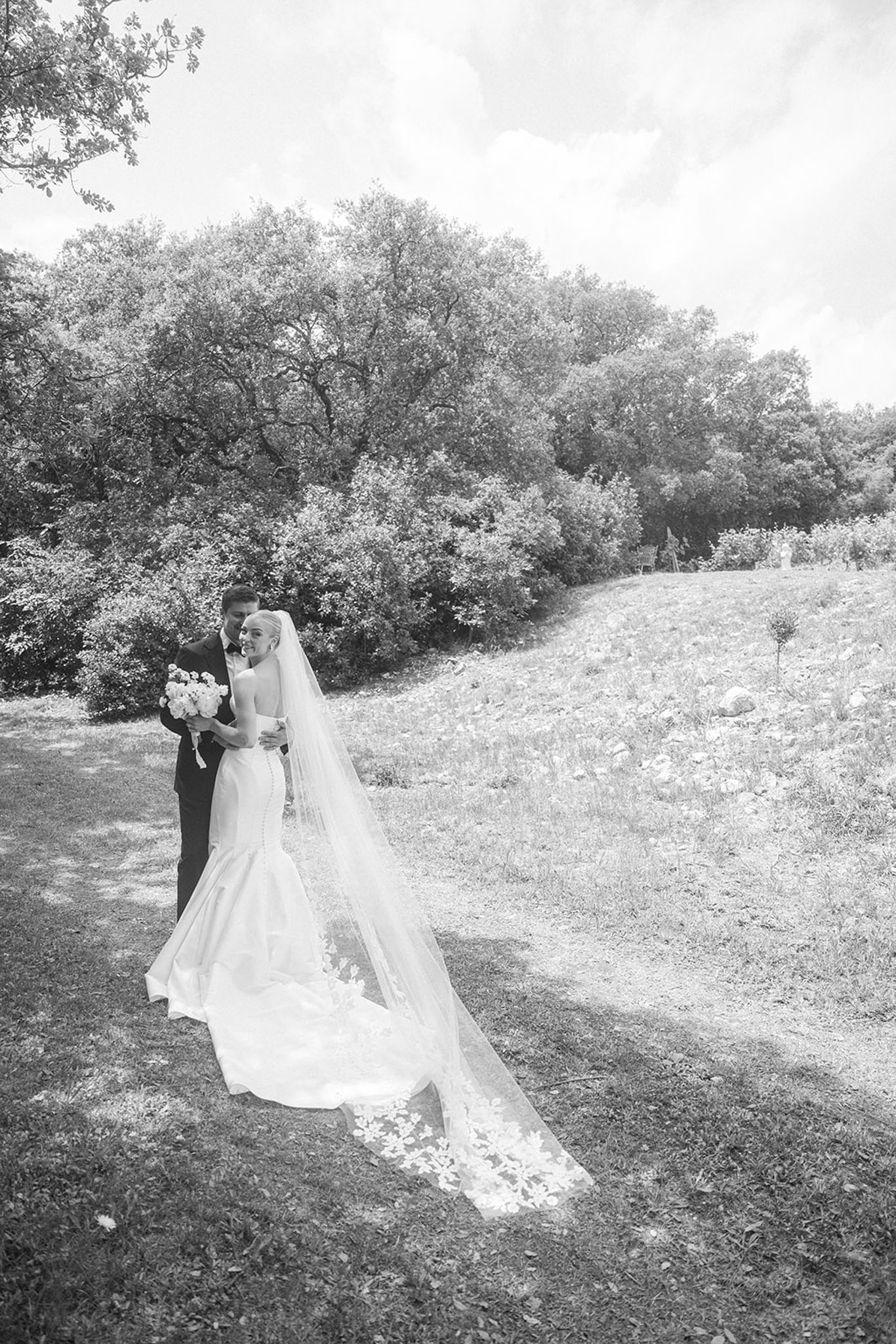 Black and white portrait of couple on hillside with bride in mermaid gown and cathedral lace veil trailing across grass