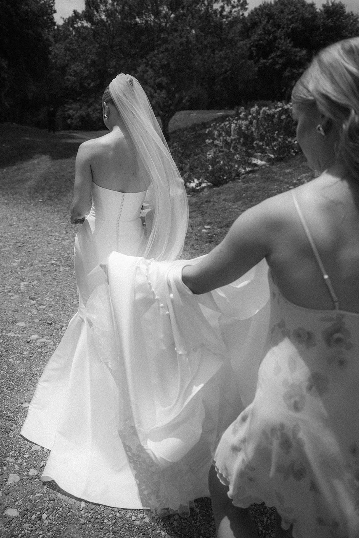 Black and white rear view of attendant carrying bride's train and veil on gravel path