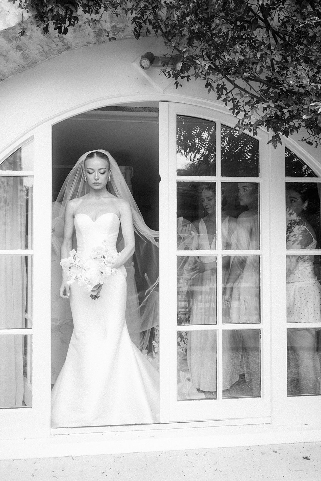 Black and white bridal portrait stepping through arched doors with bridesmaids reflected in glass