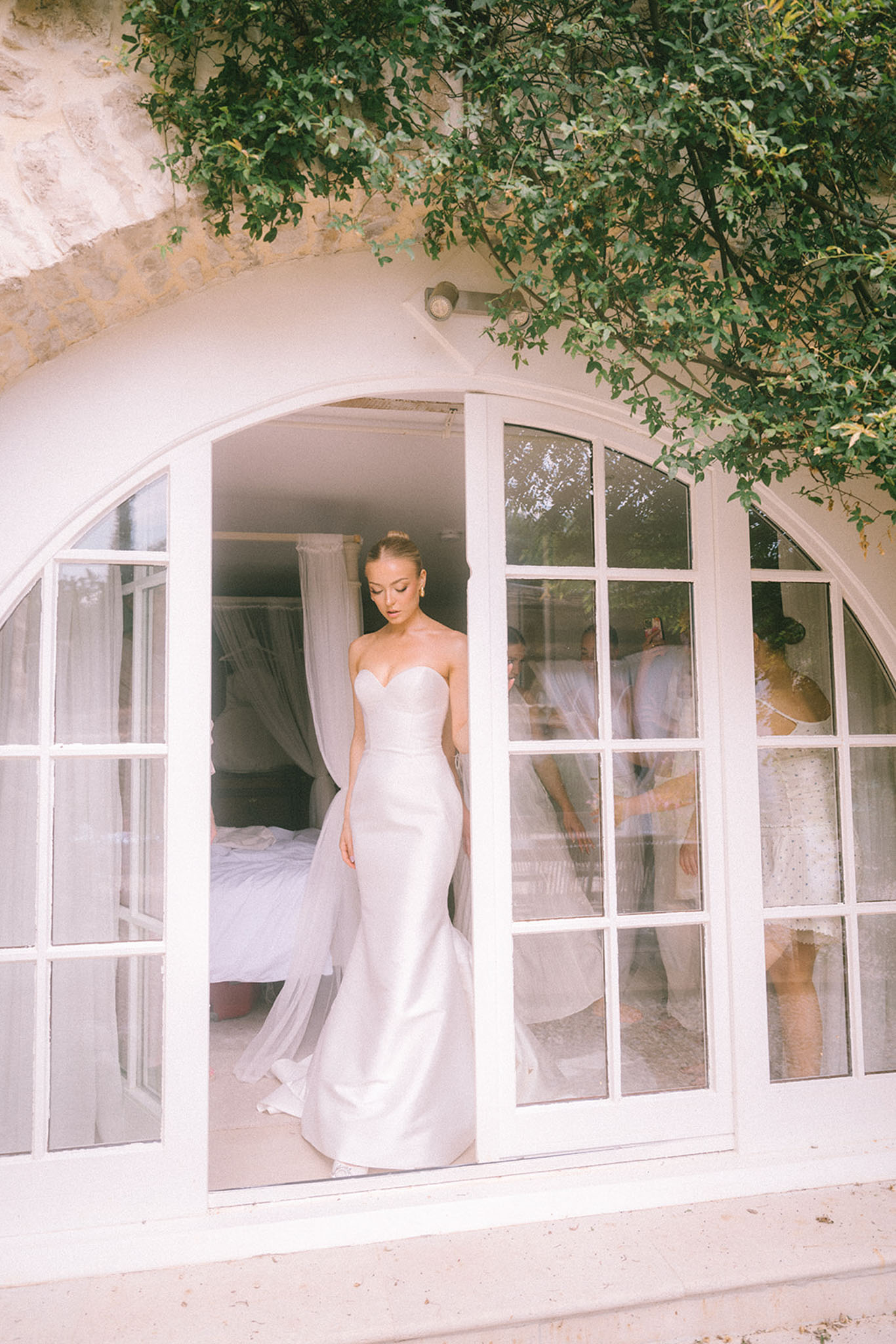Bride stepping through arched French doors in strapless sweetheart gown with tulle veil trailing behind