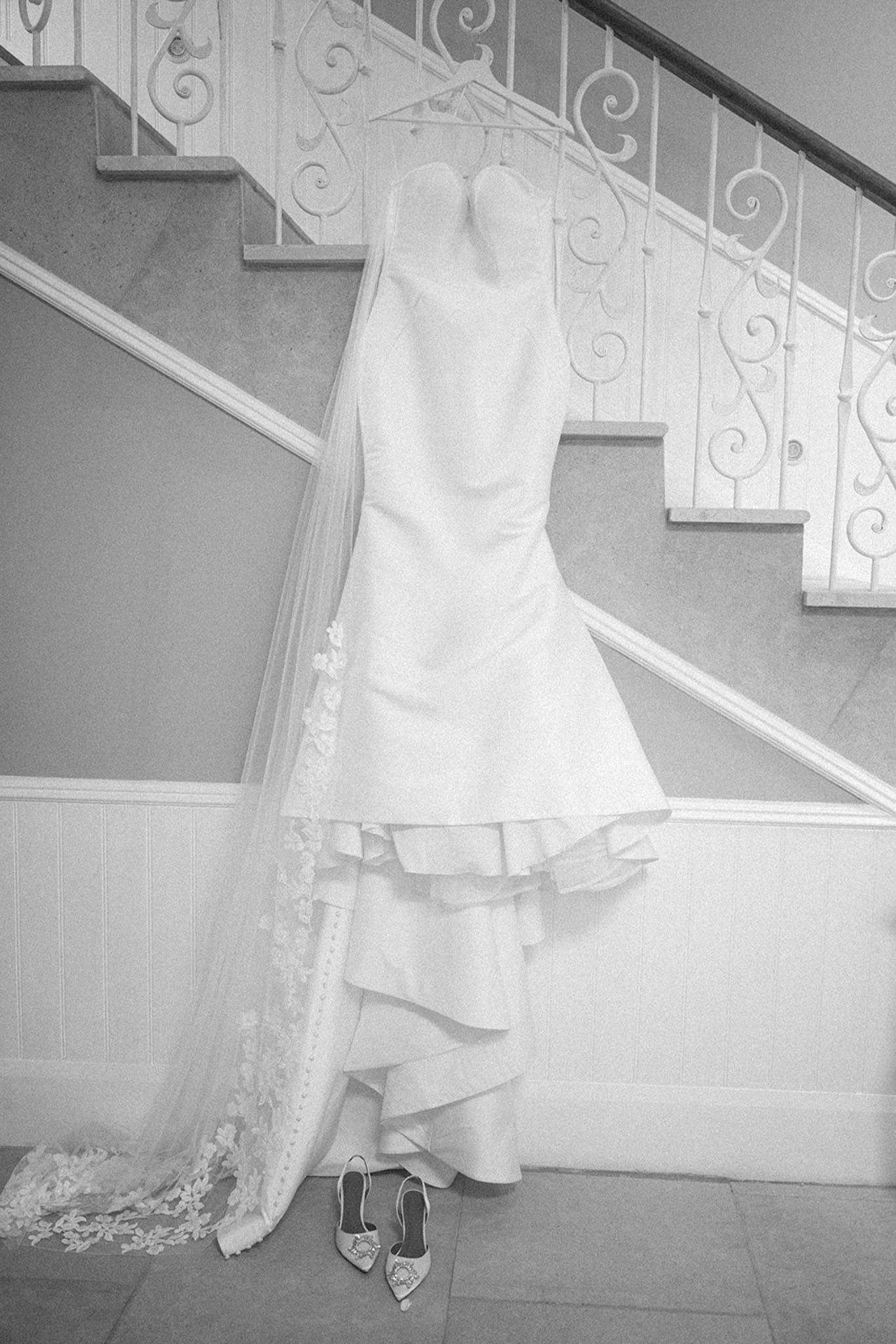 Black-and-white shot of strapless bridal gown and veil hanging on ornate iron staircase with heels below