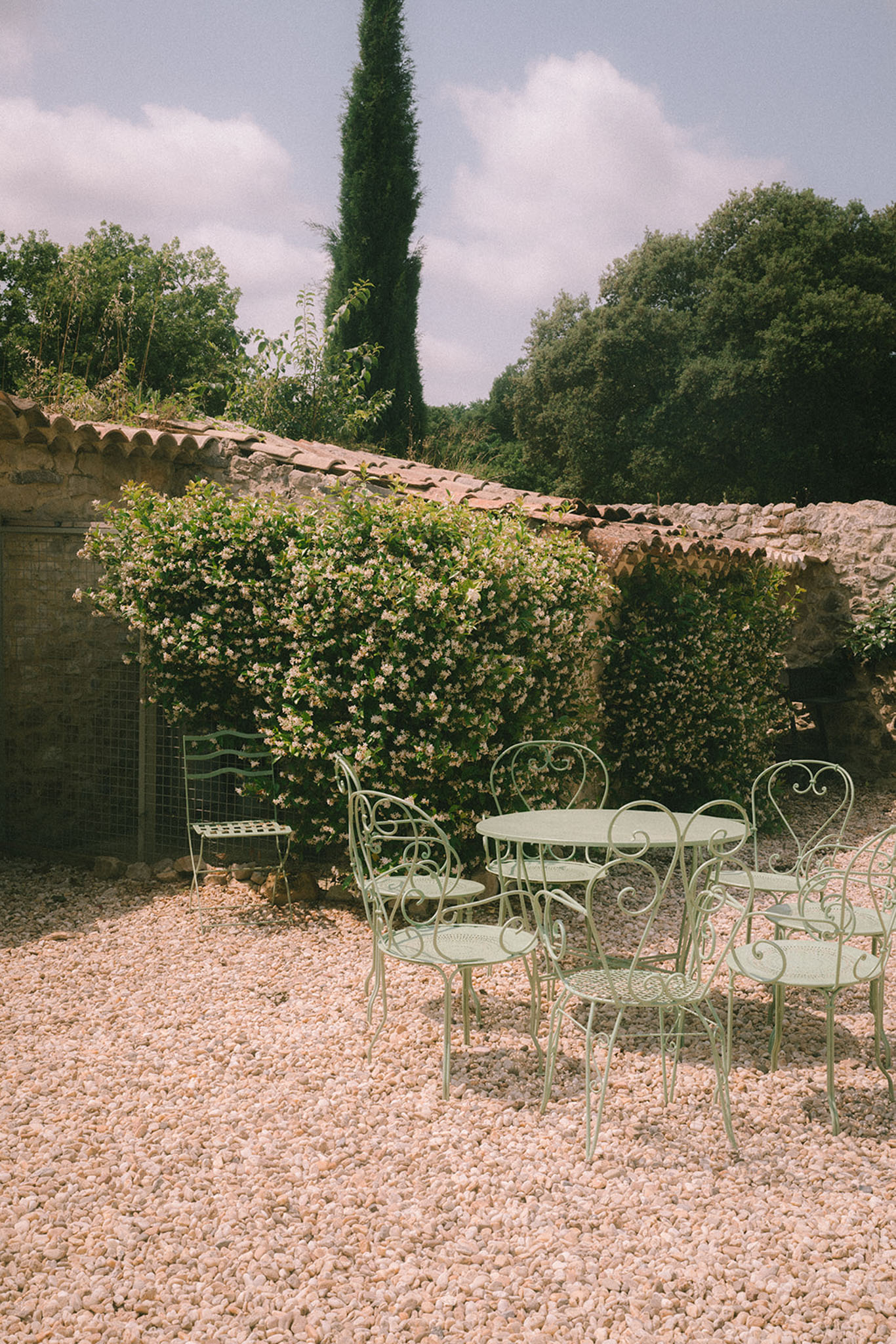 French country courtyard with sage green wrought iron garden furniture on gravel beside a stone building with climbing flo...
