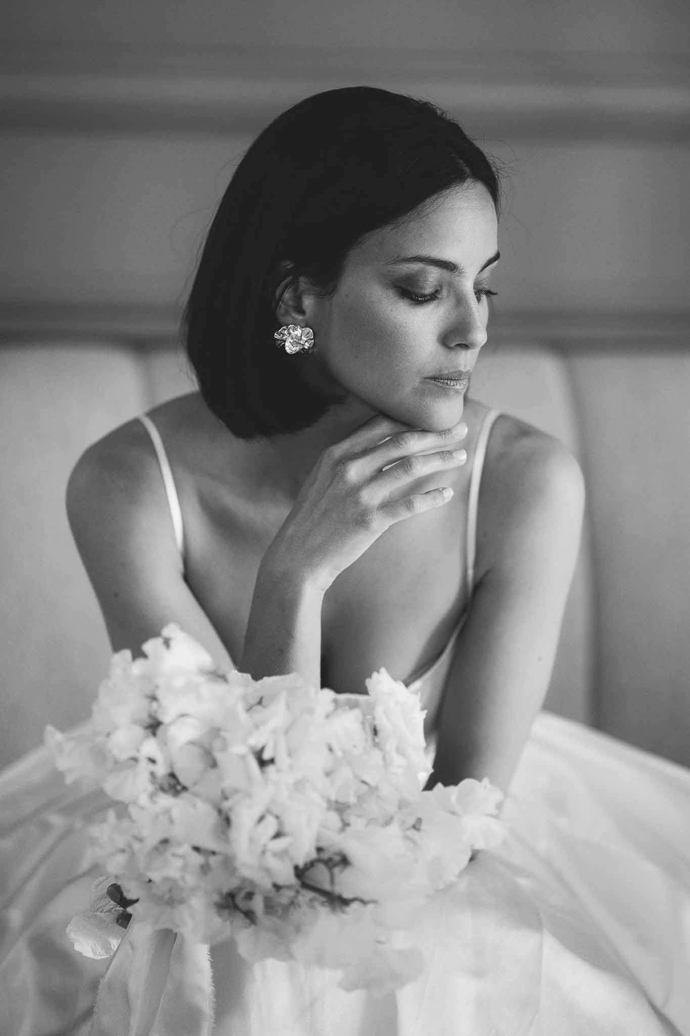 Black and white portrait of seated bride with peony bouquet in lap wearing low-cut gown and floral earrings