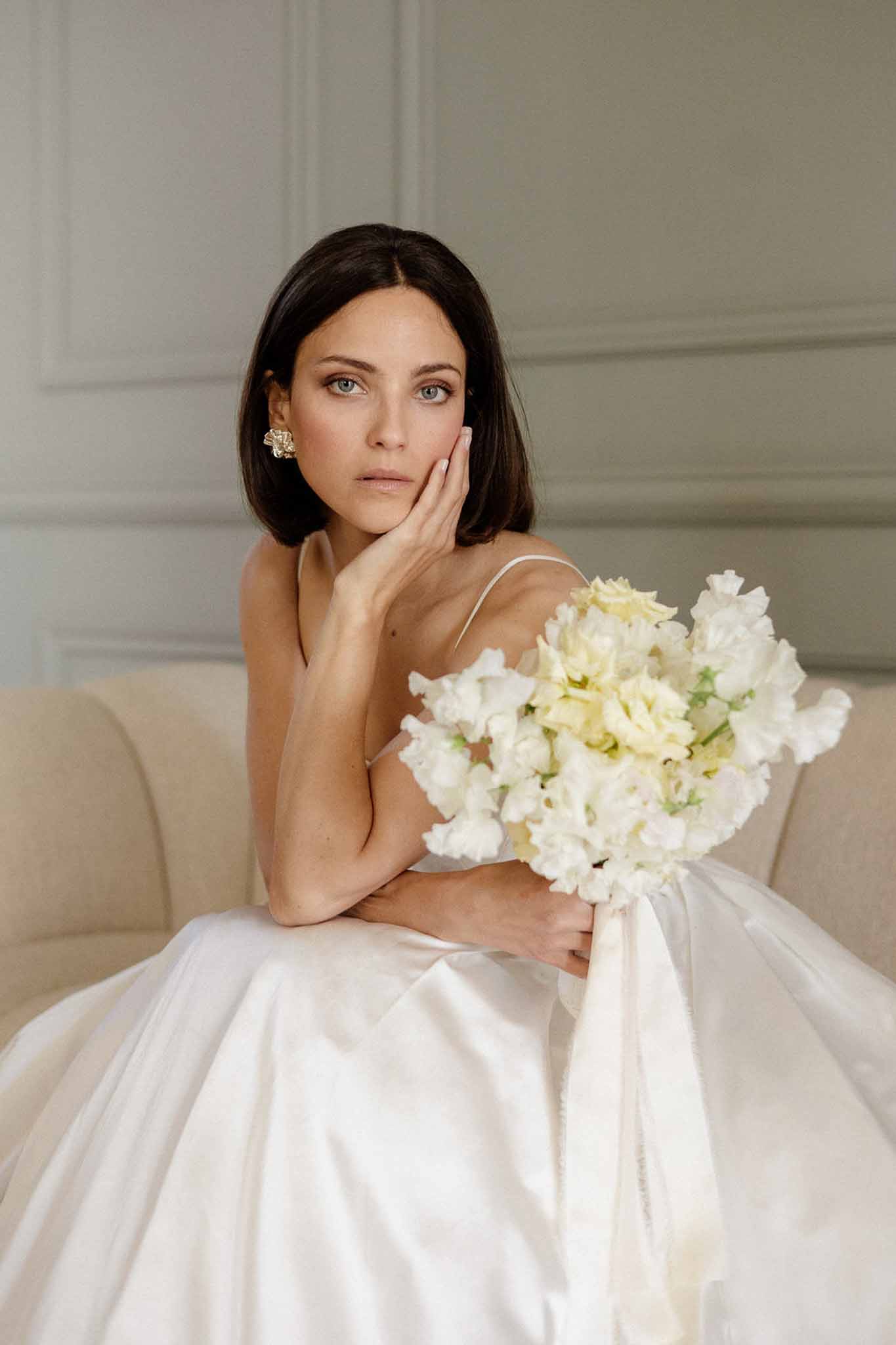 Bride with short dark hair on cream sofa holding white and yellow sweet pea bouquet with gold earring