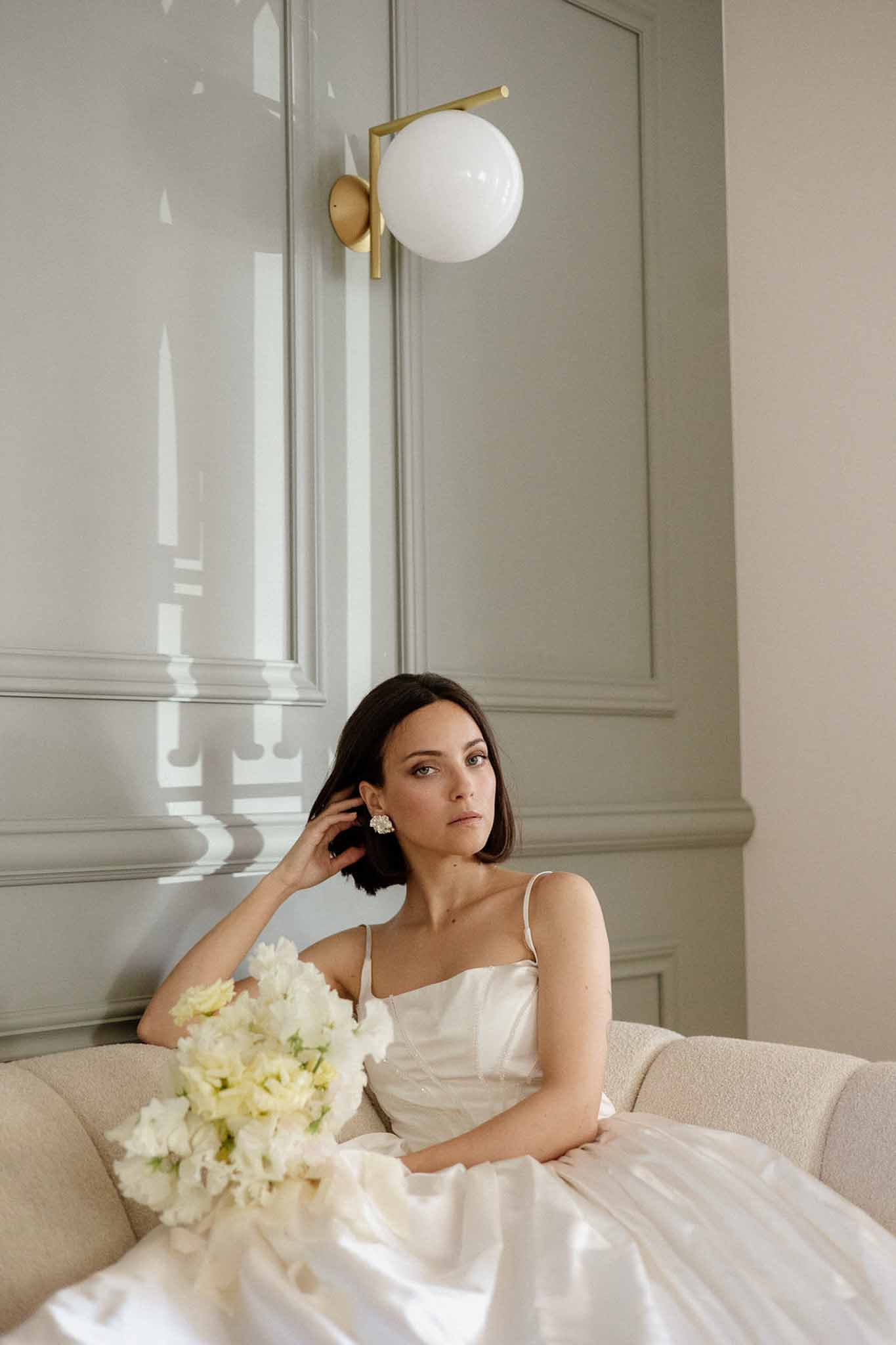 Bride in white satin slip dress with peony bouquet seated on boucle sofa in sage green panelled room