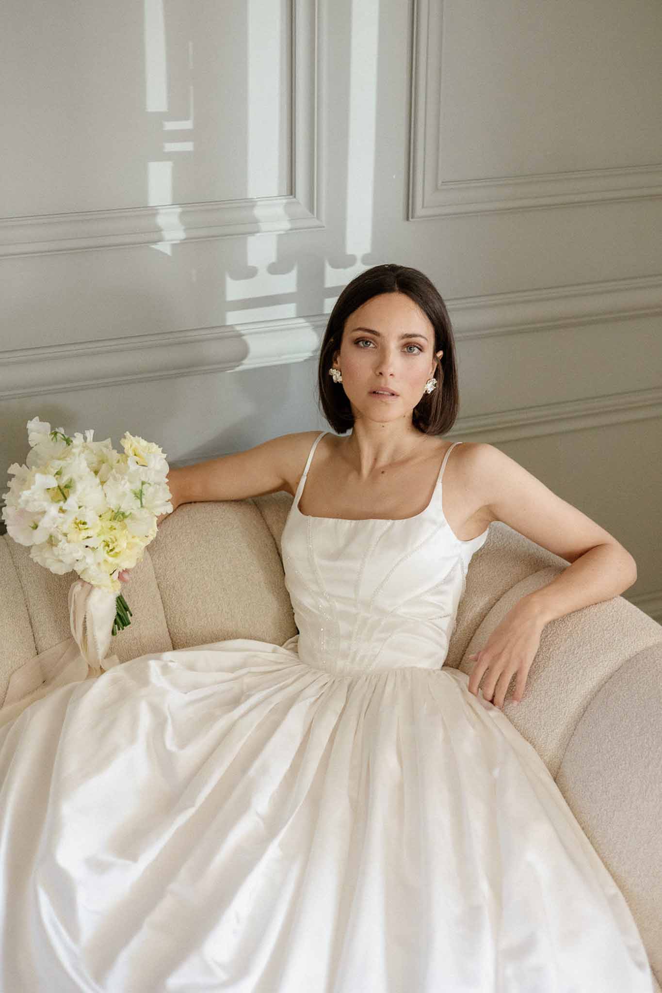 Bride in ivory satin corset gown with sweet pea bouquet and floral earrings seated on cream sofa