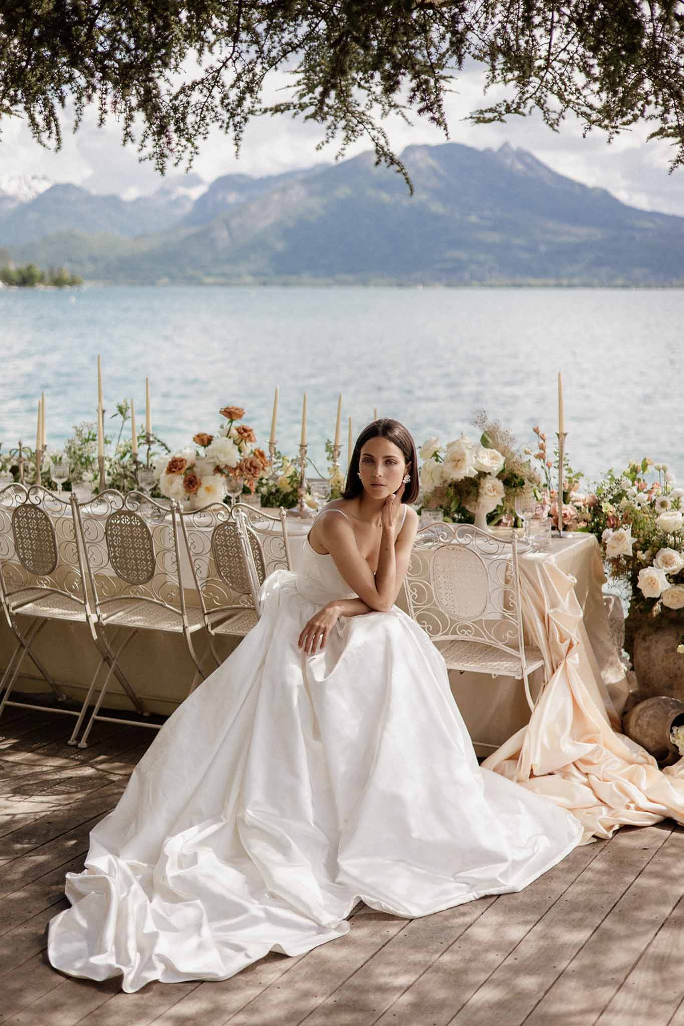 Bride in white satin ball gown seated on iron bench beside styled tablescape with roses on lakeside terrace