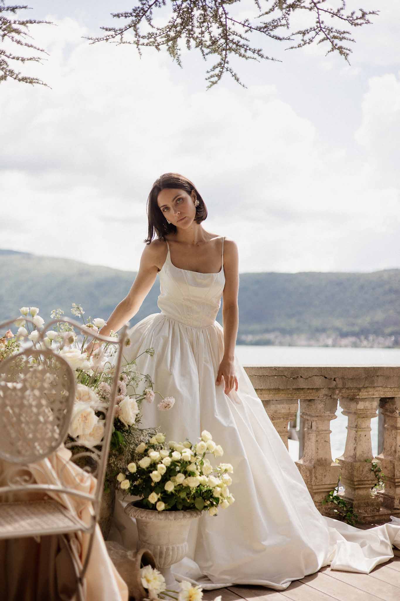 Bride in white square-neck gown beside stone urn of cream roses with lake and hill views behind