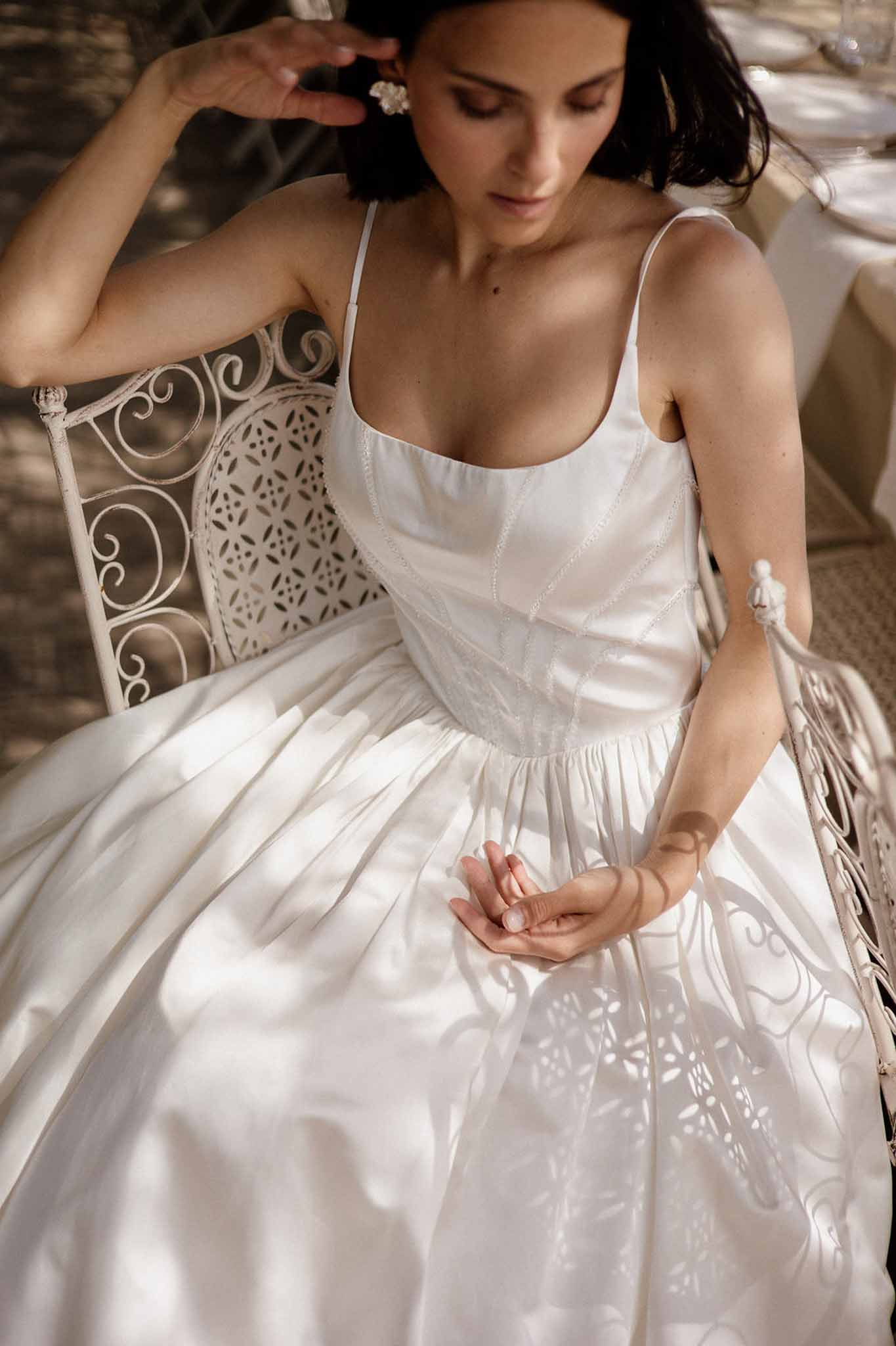 Bride seated on wrought iron chair in ivory satin gown with dappled sunlight casting shadow patterns