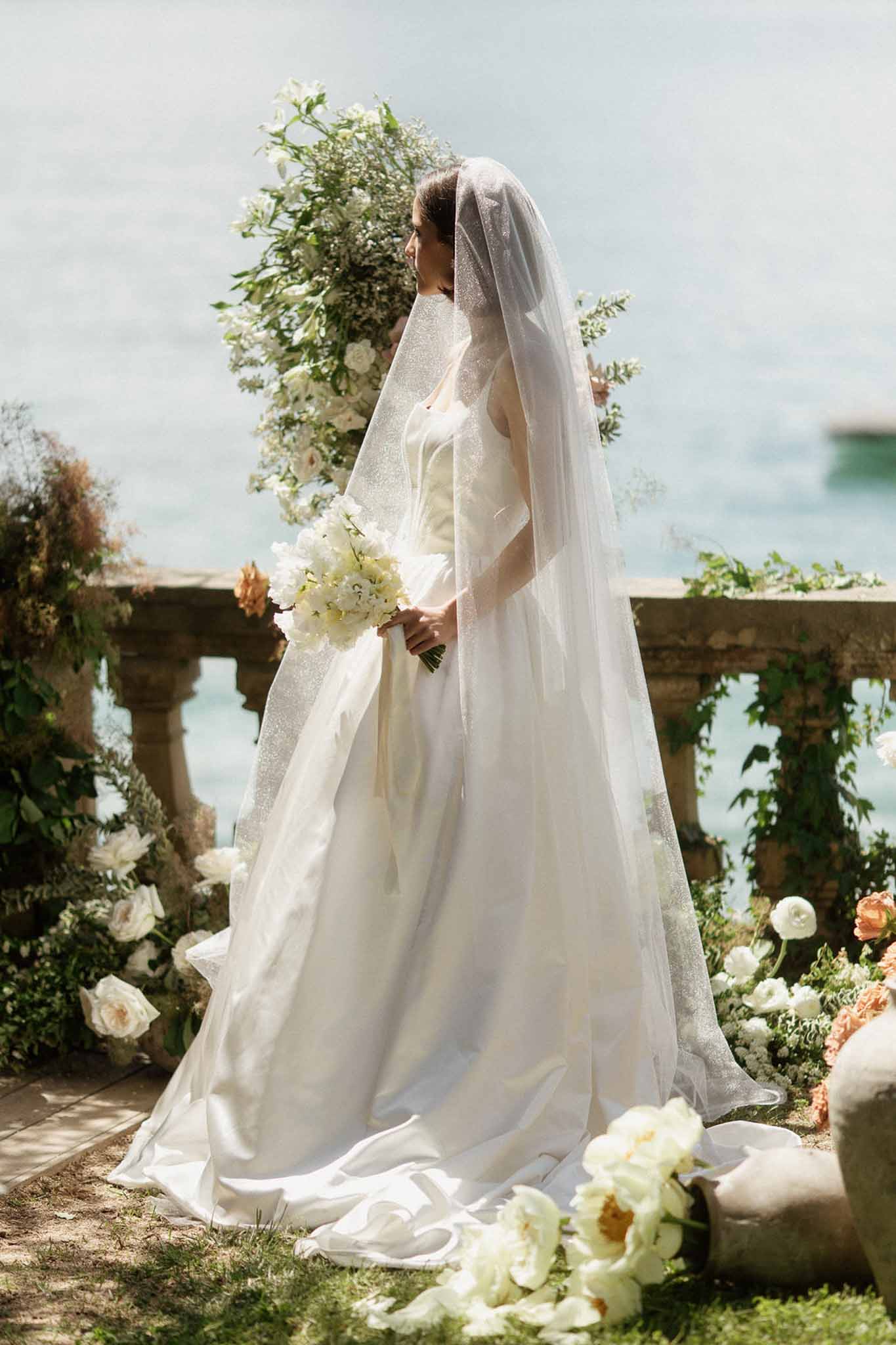 Bride in ivory ballgown with cathedral veil holding white bouquet beside lakeside stone balustrade