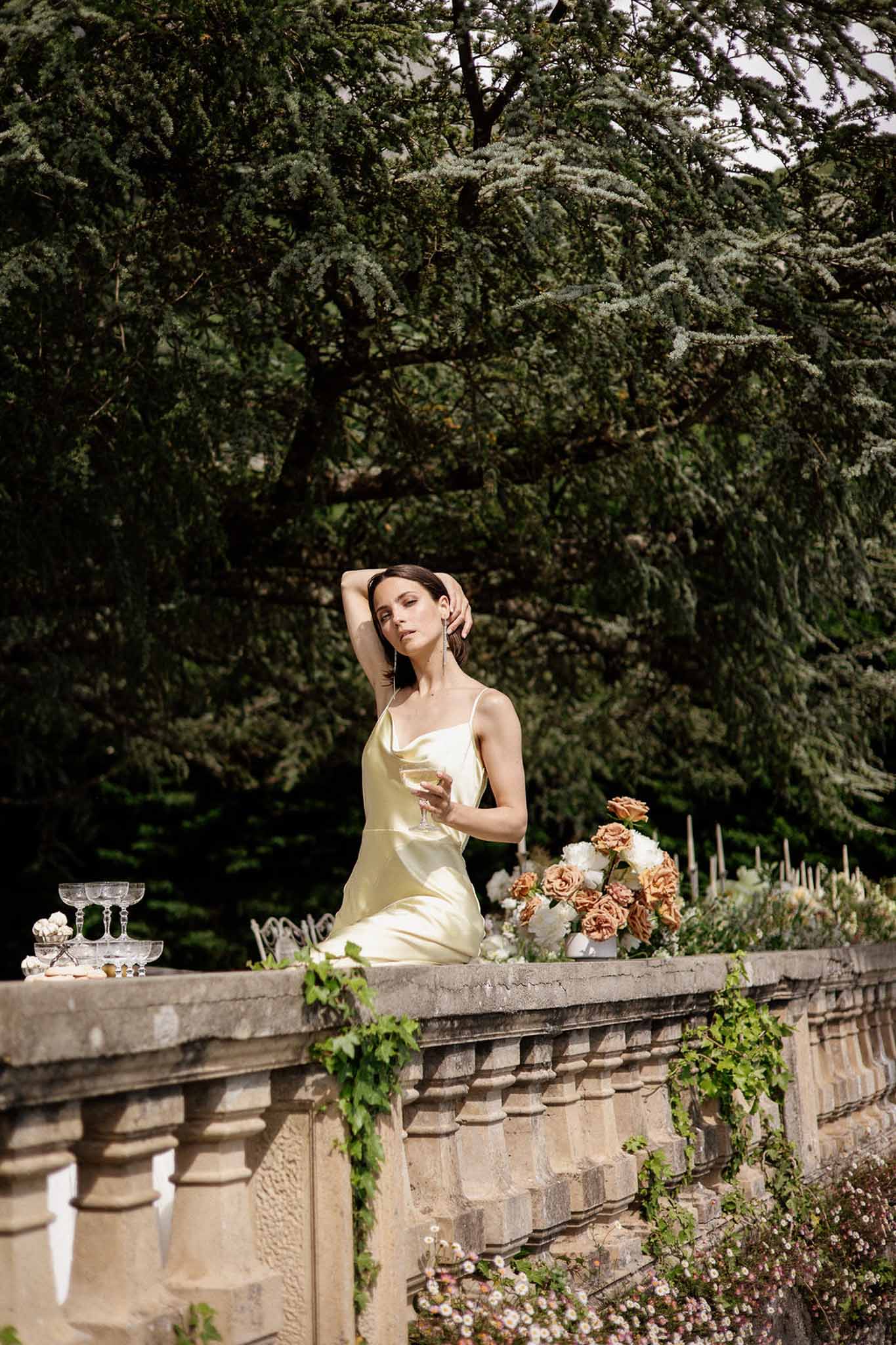 Woman in yellow silk slip dress with champagne coupe beside orange rose and ivory peony arrangement
