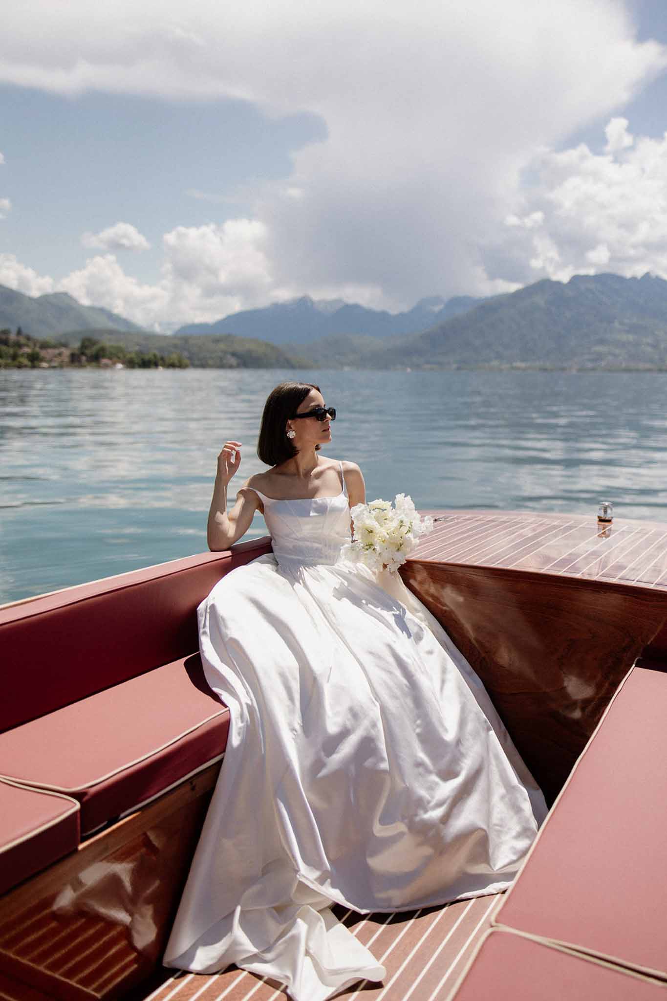 Bride in off-shoulder ball gown with sunglasses holding peony bouquet on wooden boat on alpine lake