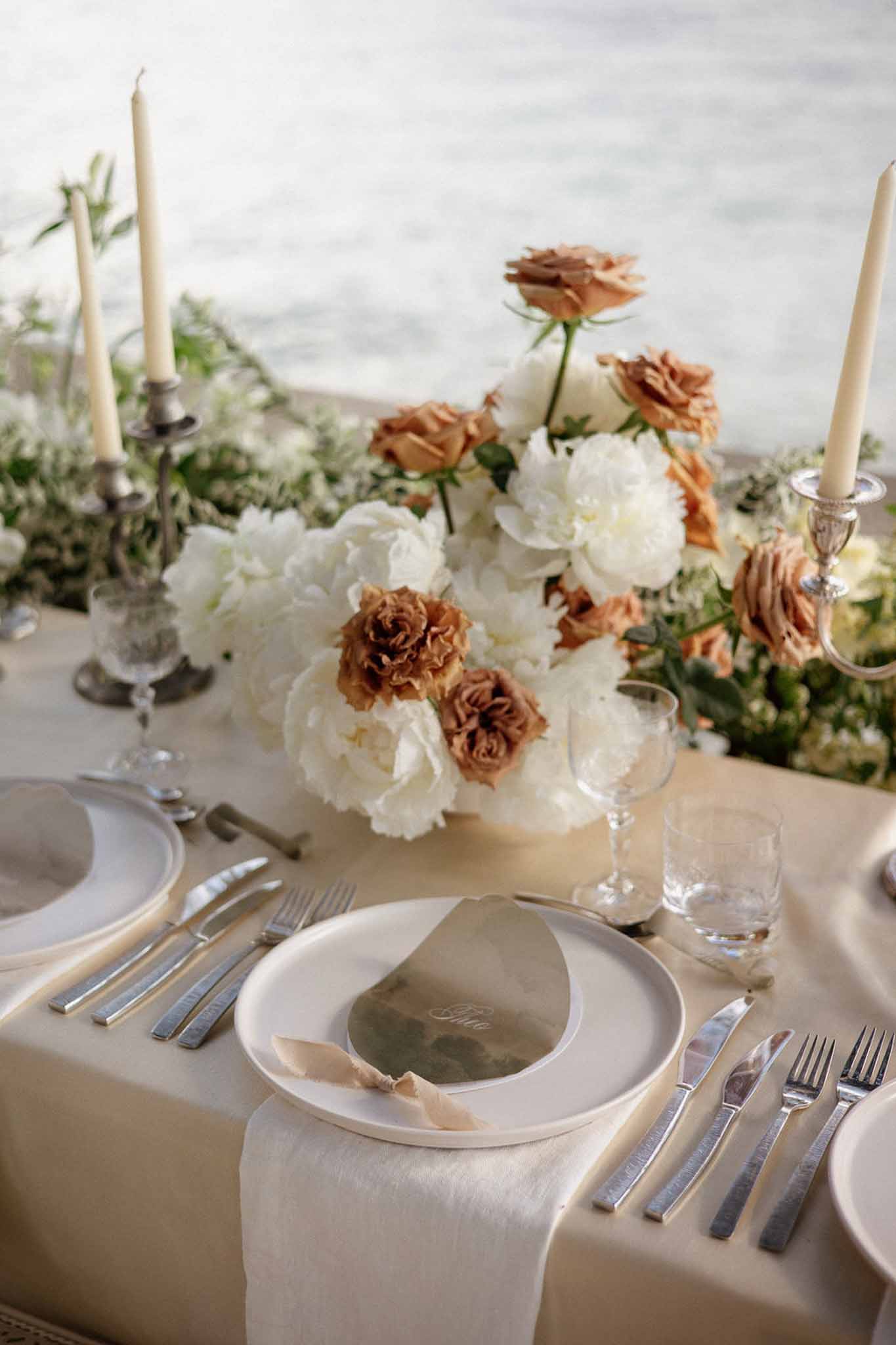 Lakeside table with white peonies brown roses greenery garland and metallic place cards on sand linen