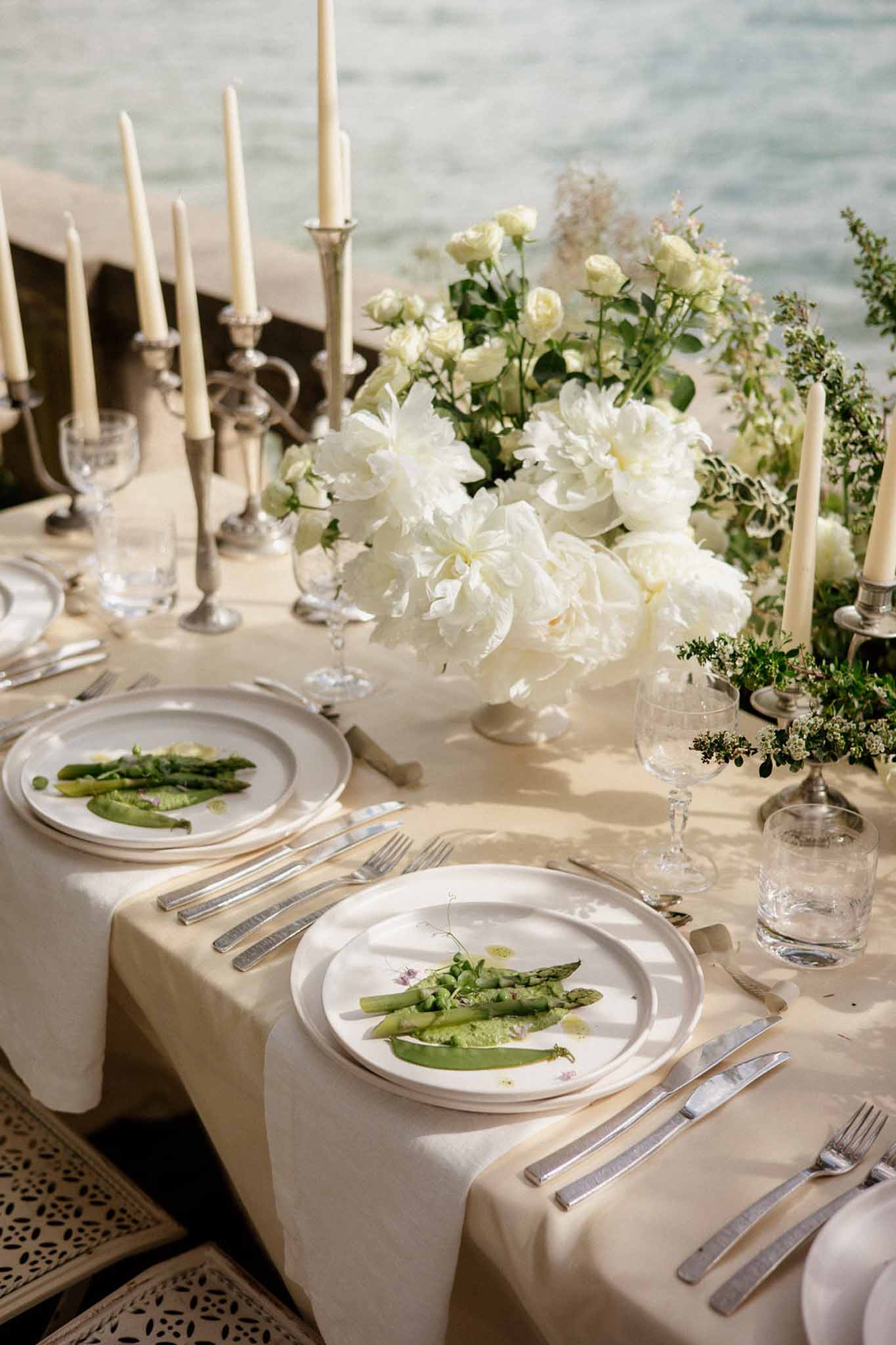 Close-up of waterside reception table with white peony centerpiece, silver candelabras, and plated asparagus starter
