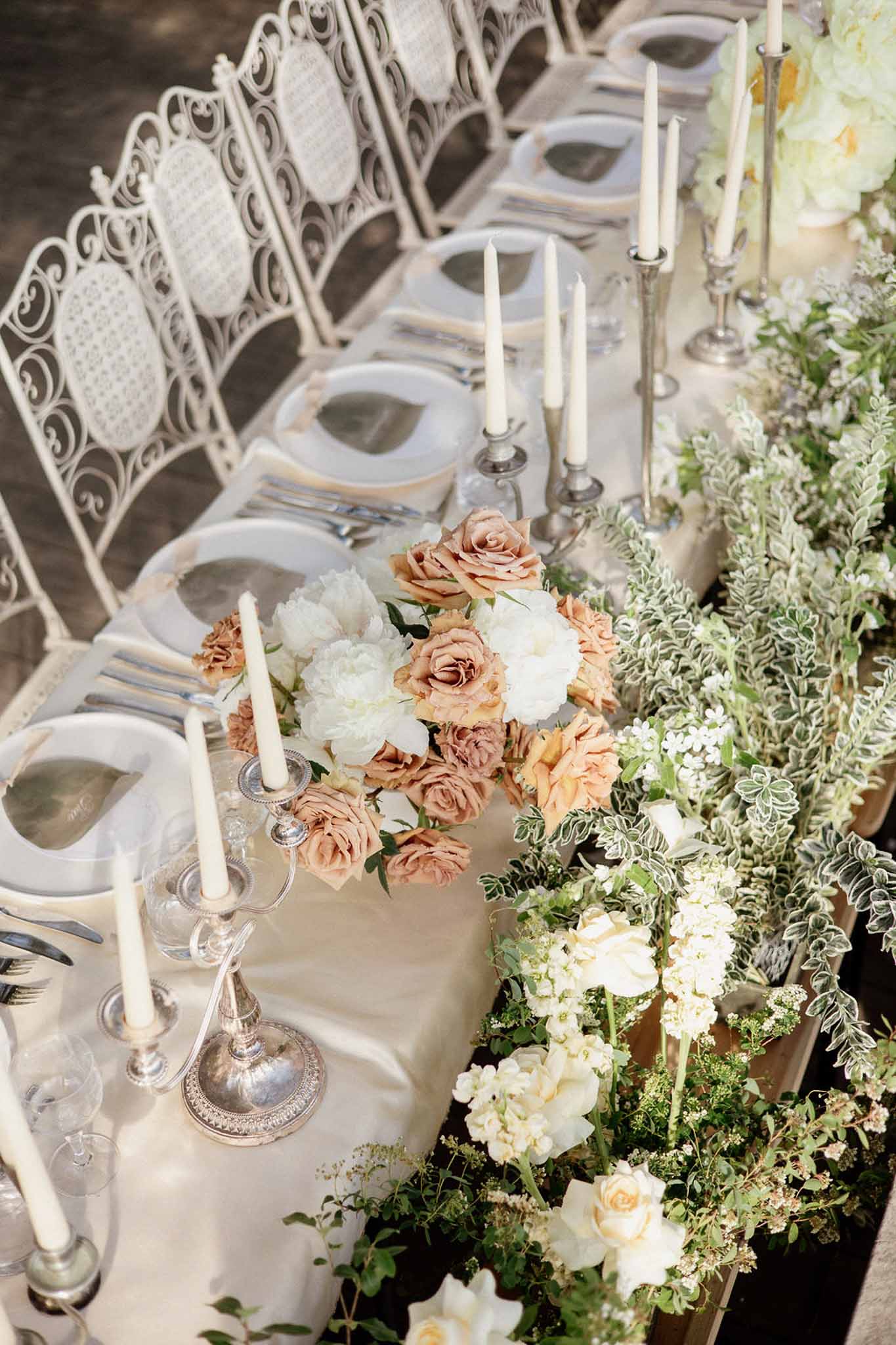 Outdoor reception table with cream runner, dusty mauve and caramel rose centerpiece, ivory taper candles, and white wrough...